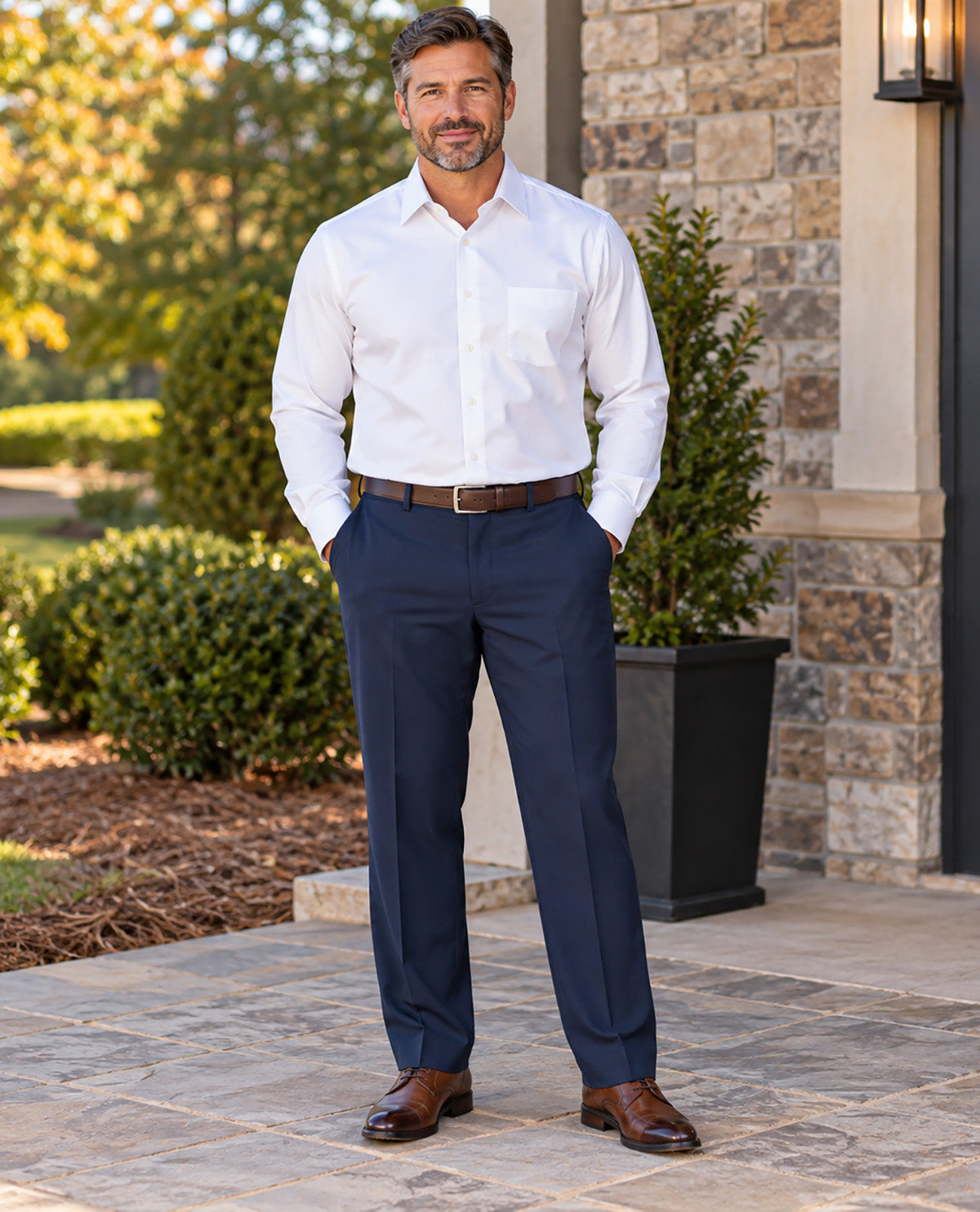 A man stands outdoors on a stone walkway in front of a house, wearing a white long-sleeve dress shirt, navy dress pants, a brown belt, and brown dress shoes.