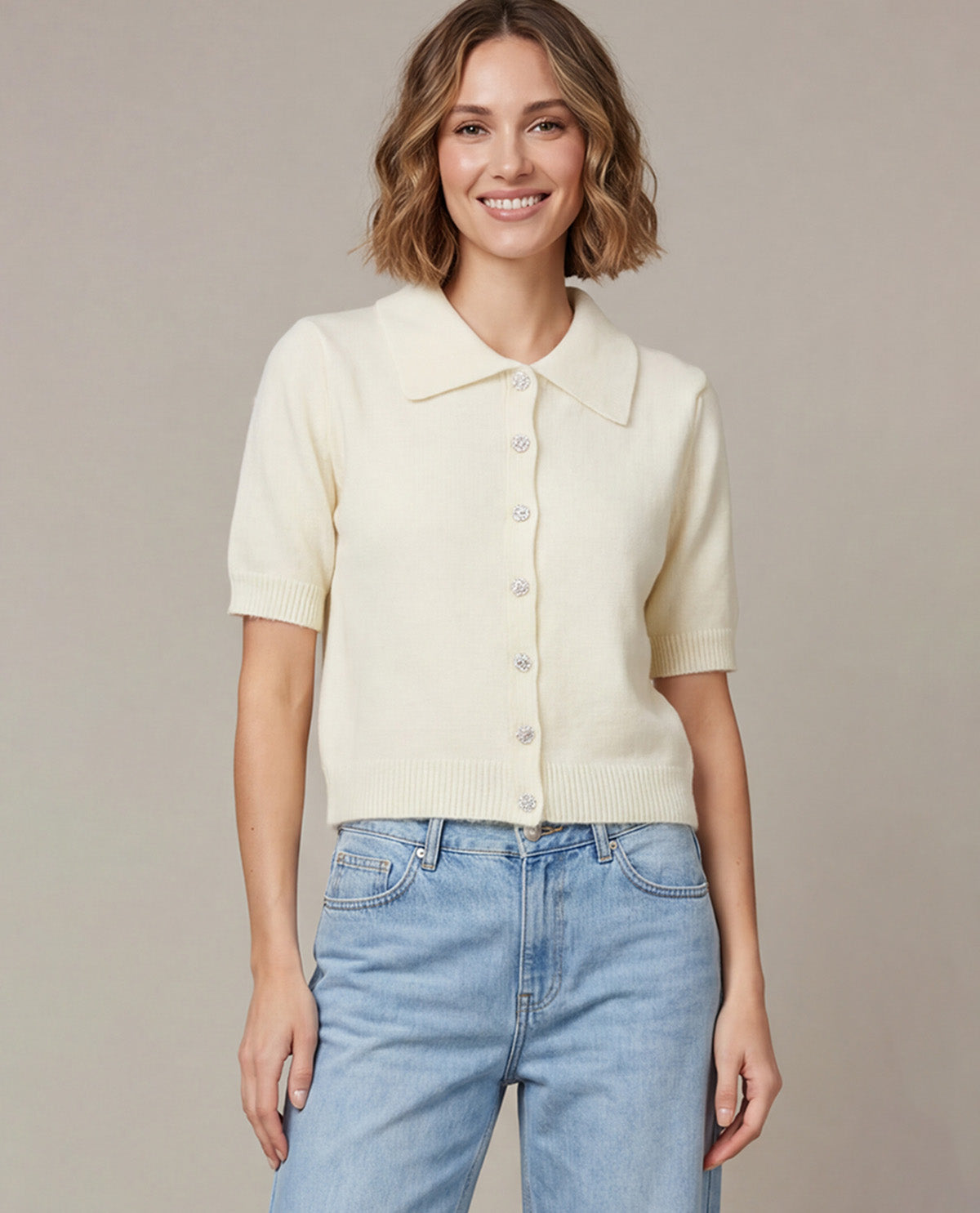 A woman wearing a cream-colored short-sleeve knit button-up cardigan with a collar, paired with light blue jeans, standing against a neutral beige studio background.