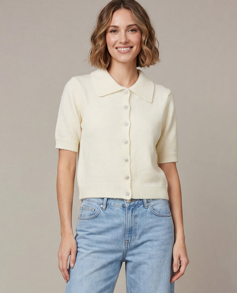 A woman wearing a cream-colored short-sleeve knit button-up cardigan with a collar, paired with light blue jeans, standing against a neutral beige studio background.