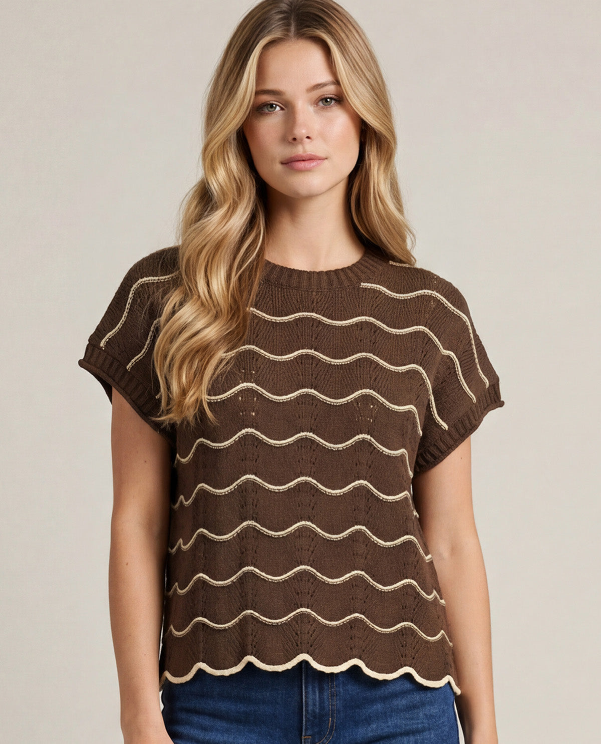 A woman wearing a brown short-sleeve knit top with horizontal wavy beige stripes, paired with blue jeans, against a plain beige studio background.