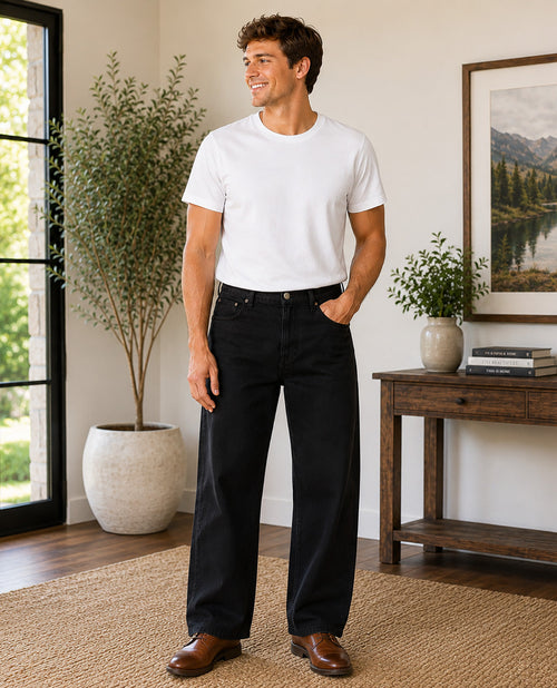 A man wearing a white crewneck t-shirt and black wide-leg jeans, standing in a bright, modern room with a wooden console table, potted plants, and a framed landscape print.