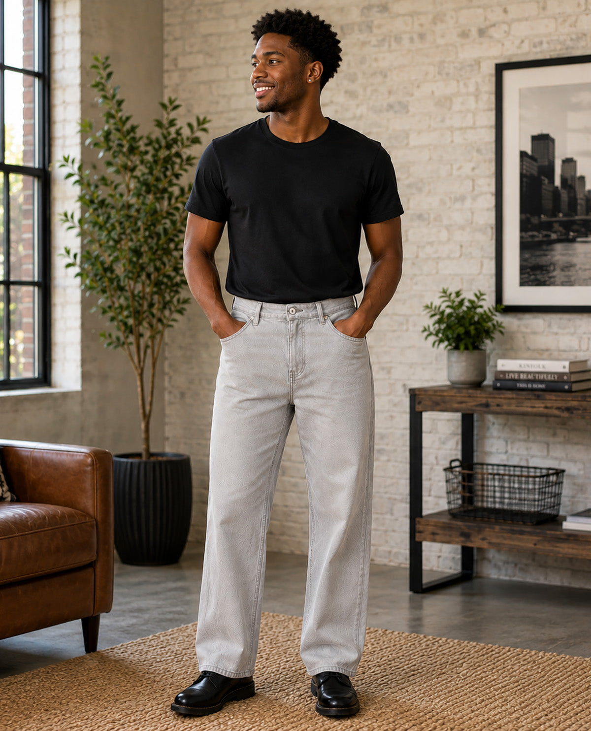A man wearing a black crewneck t-shirt and light gray straight-leg jeans, standing in a modern loft-style room with a brick wall, potted plants, and a brown leather armchair.
