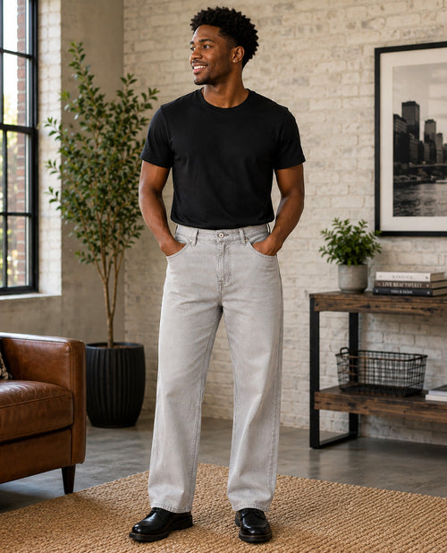 A man wearing a black crewneck t-shirt and light gray straight-leg jeans, standing in a modern loft-style room with a brick wall, potted plants, and a brown leather armchair.