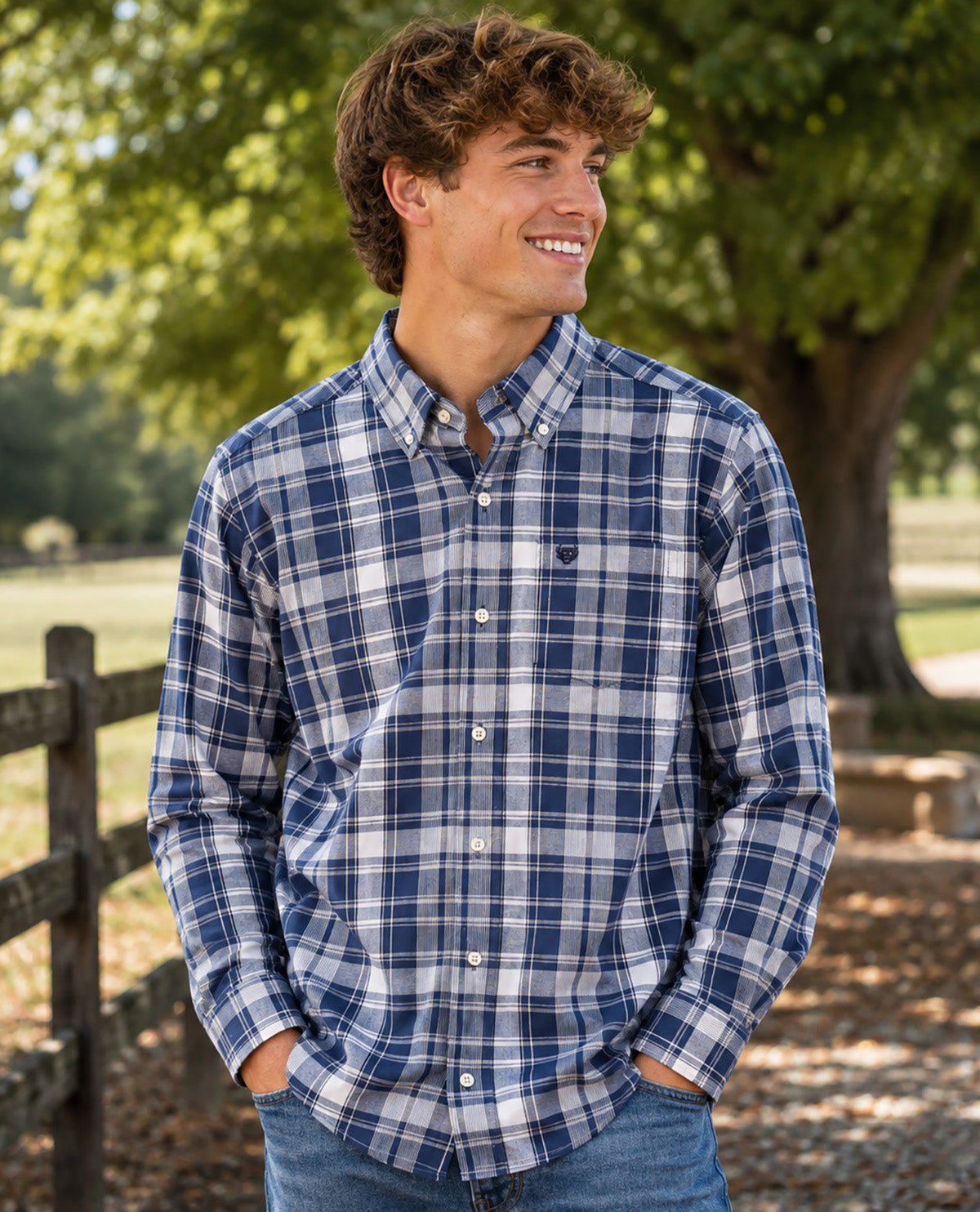 A smiling young man outdoors wearing a blue and white plaid button-down shirt with jeans, standing in front of a tree and wooden fence.