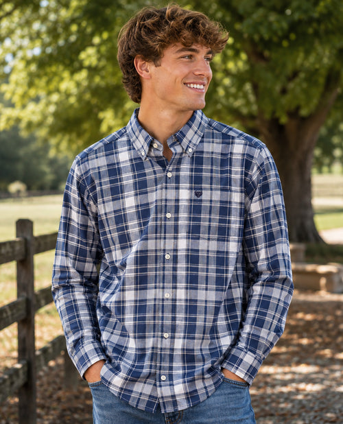 A smiling young man outdoors wearing a blue and white plaid button-down shirt with jeans, standing in front of a tree and wooden fence.