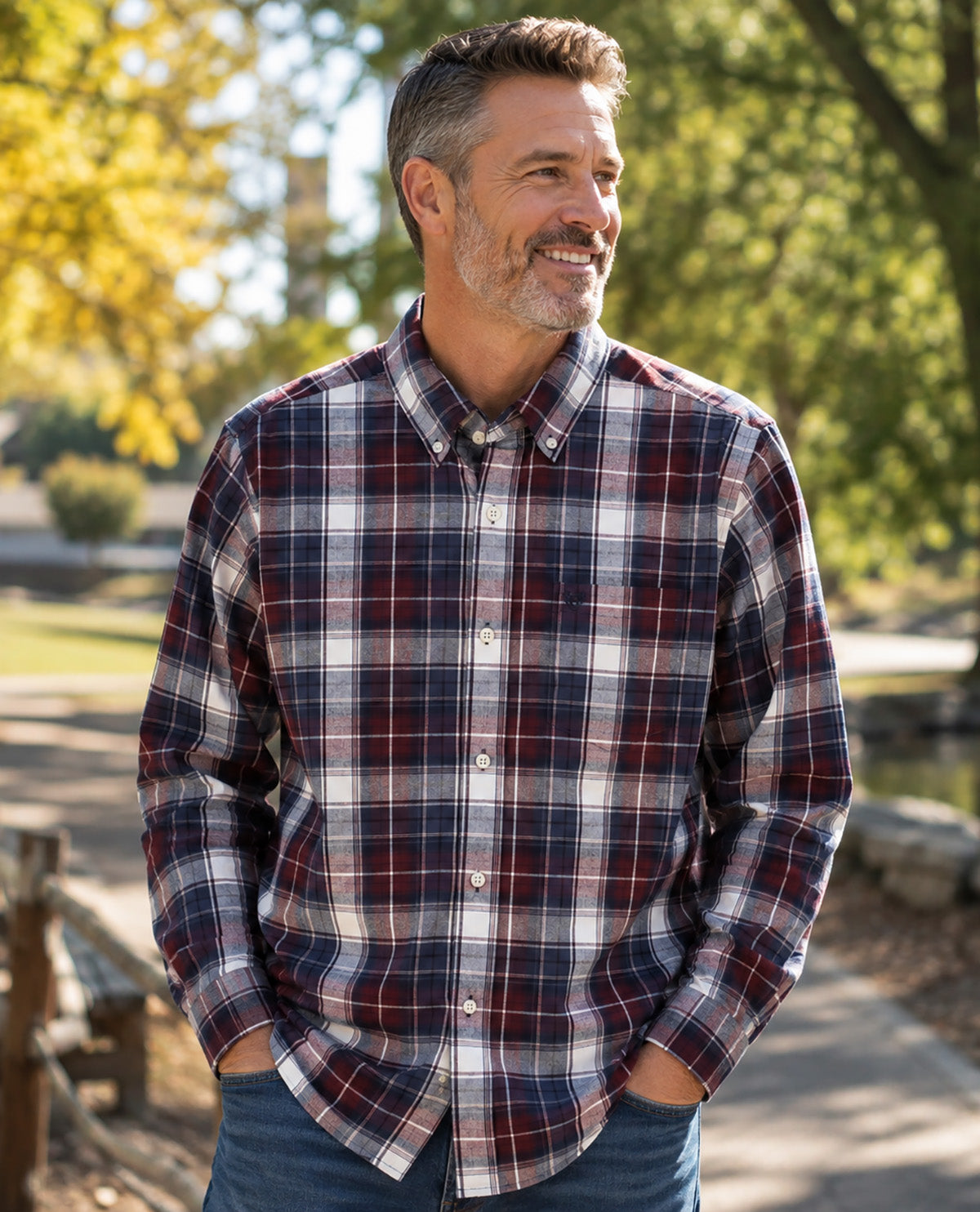 A smiling man wearing a long-sleeve button-down plaid shirt in navy, red, and white, paired with blue jeans, standing outdoors in a park with trees in the background.