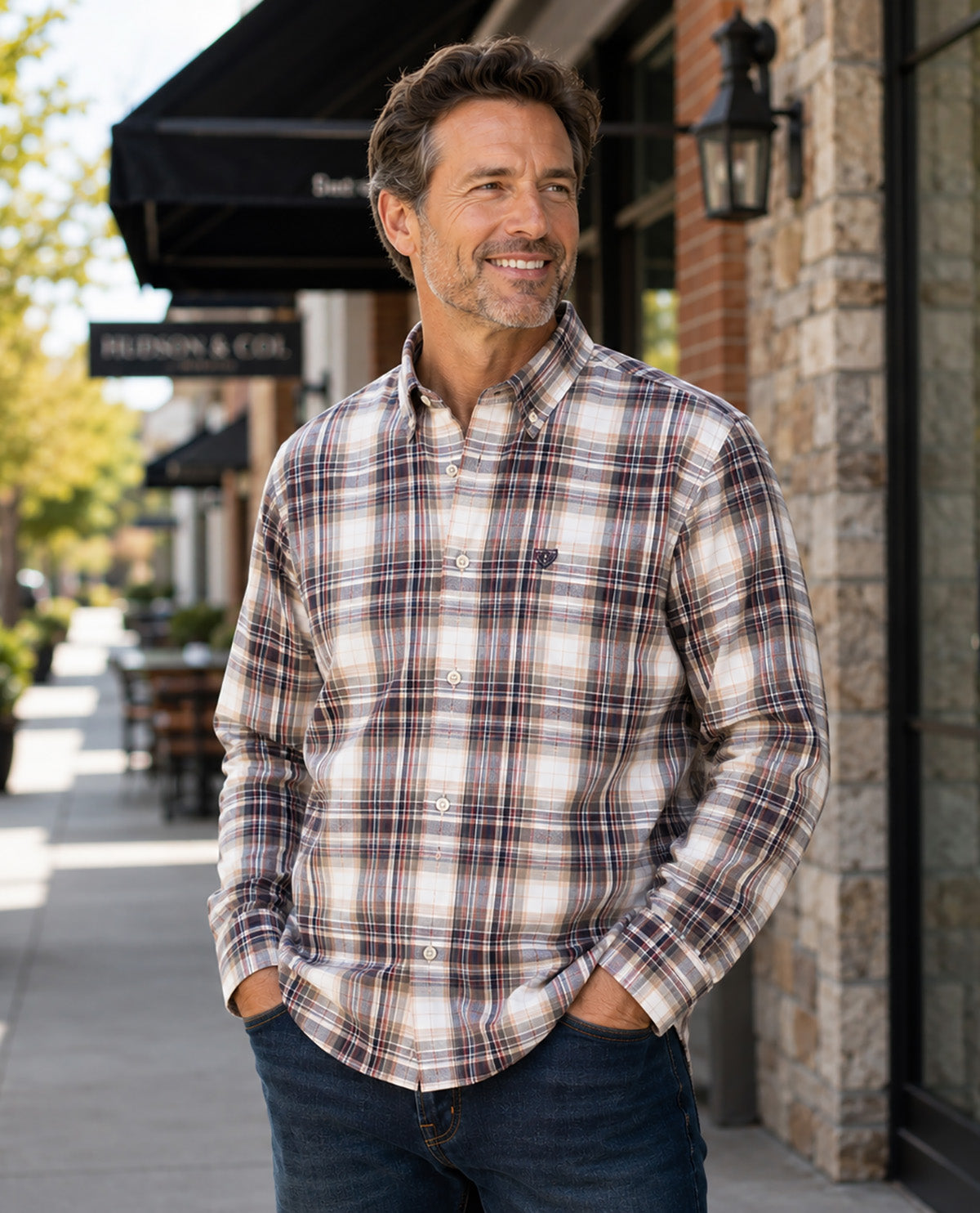 A man wearing a long-sleeve button-down plaid shirt in brown, beige, and white tones, paired with dark blue jeans, standing outdoors on a sidewalk in front of a brick building.