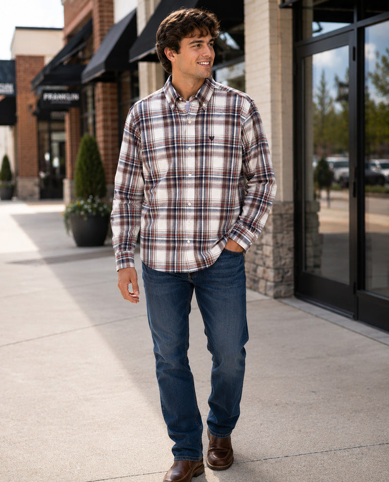 A man wearing a long-sleeve button-down plaid flannel shirt in brown, beige, and white, paired with blue jeans and brown leather shoes, walking on a sunny outdoor sidewalk.