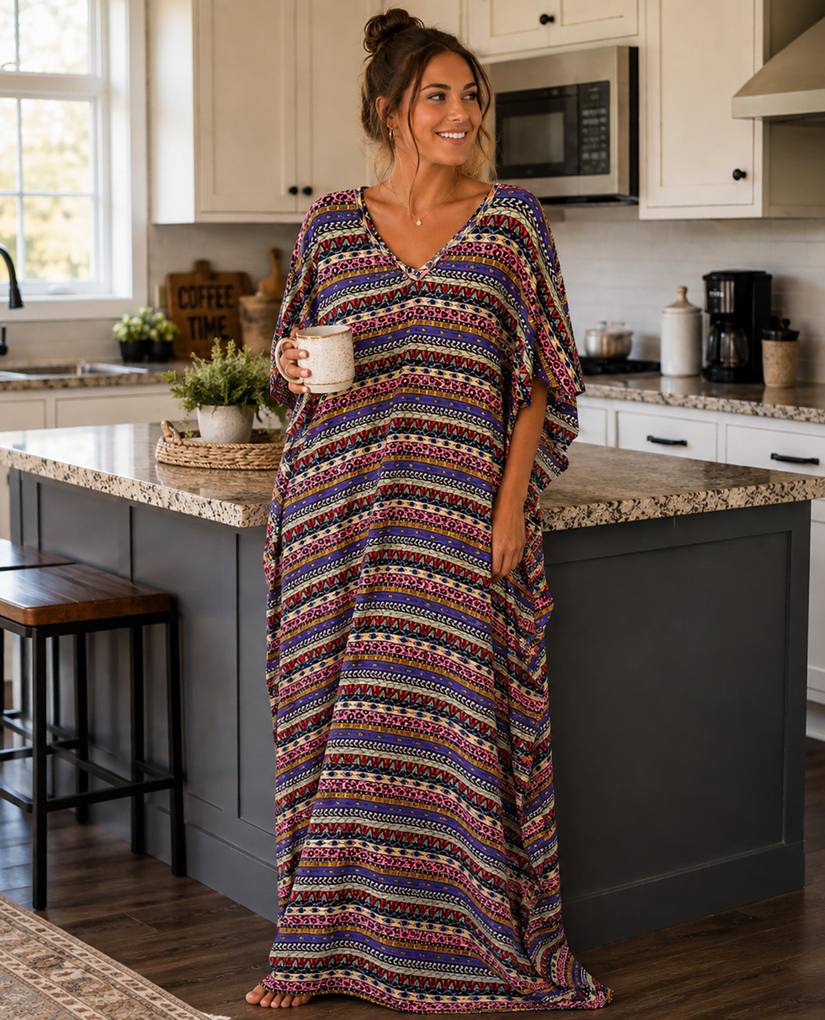 A woman wearing a long, loose kaftan dress with a colorful geometric pattern stands in a bright kitchen, holding a white mug.