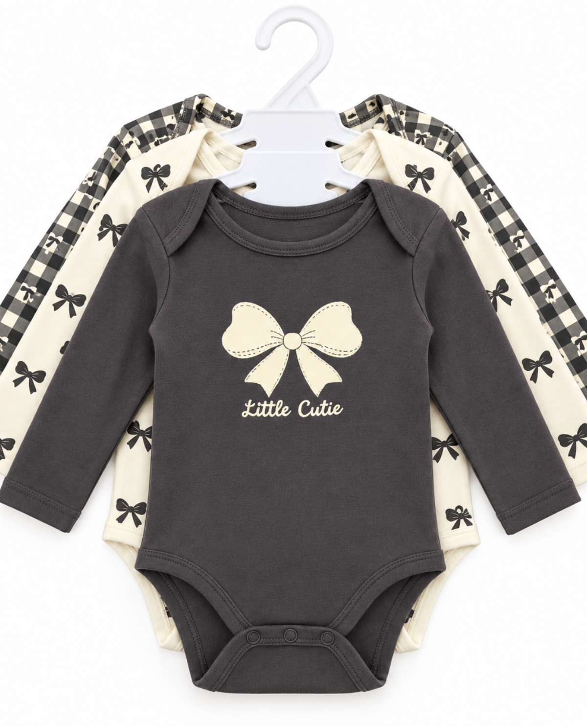 A set of three baby long-sleeve bodysuits hanging on a white plastic hanger against a white background. The front bodysuit is dark charcoal gray with a cream bow graphic and 'Little Cutie' text.