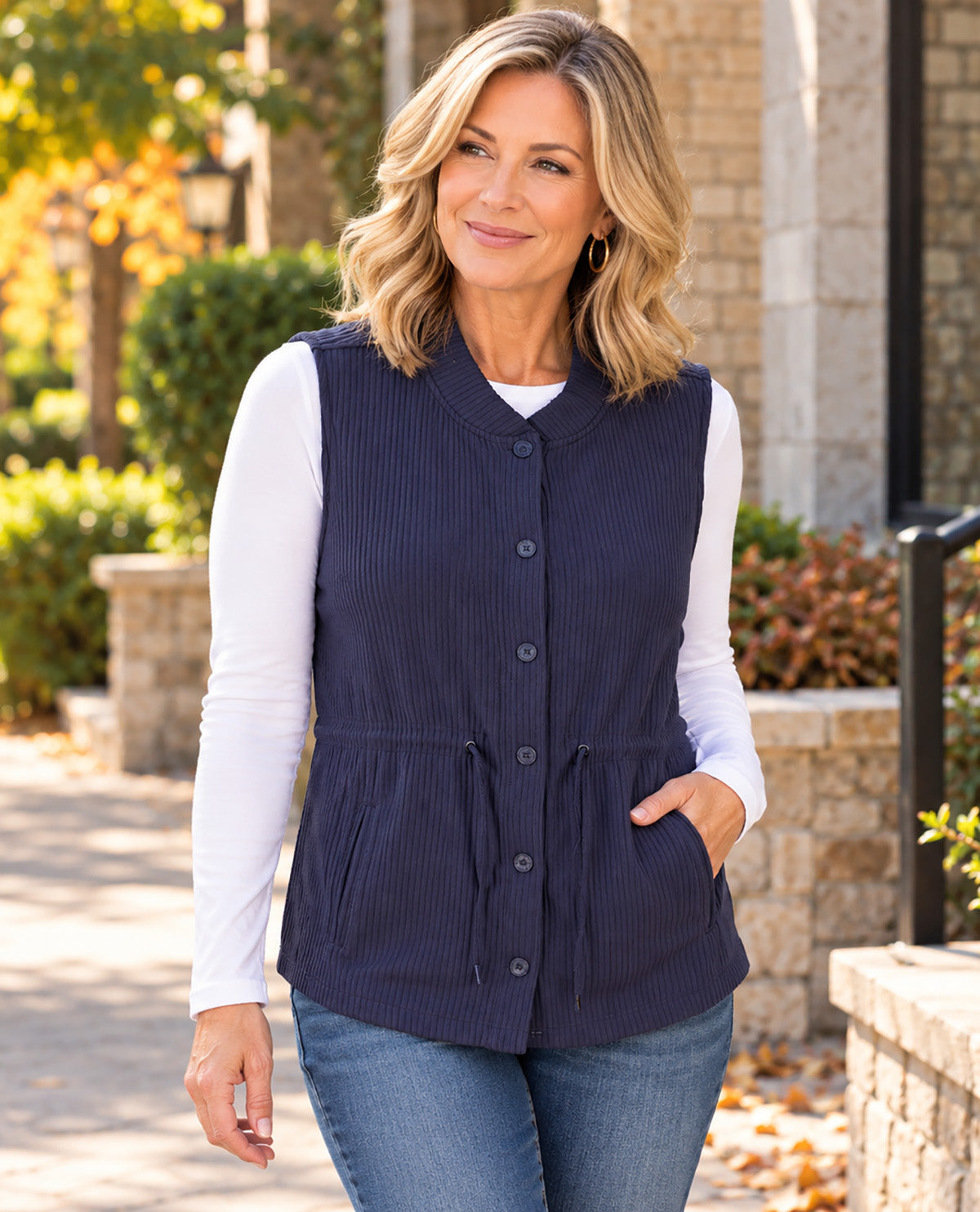 Missy model wearing a navy vest with a white long sleeve underneath paired with jeans with an outdoor background