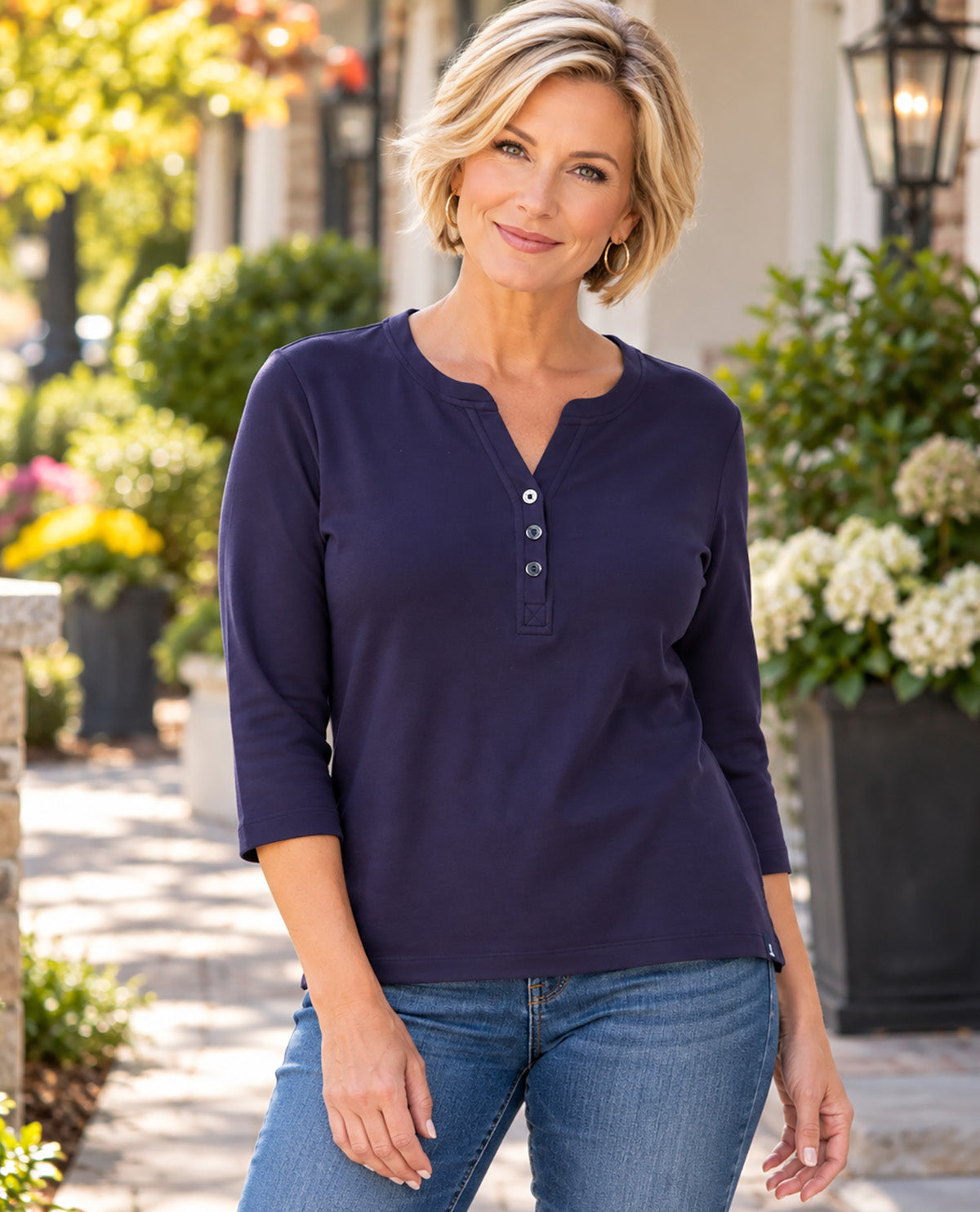 A woman wearing a navy blue henley top with three-quarter sleeves and blue jeans, standing outdoors in front of a blurred garden and house background.