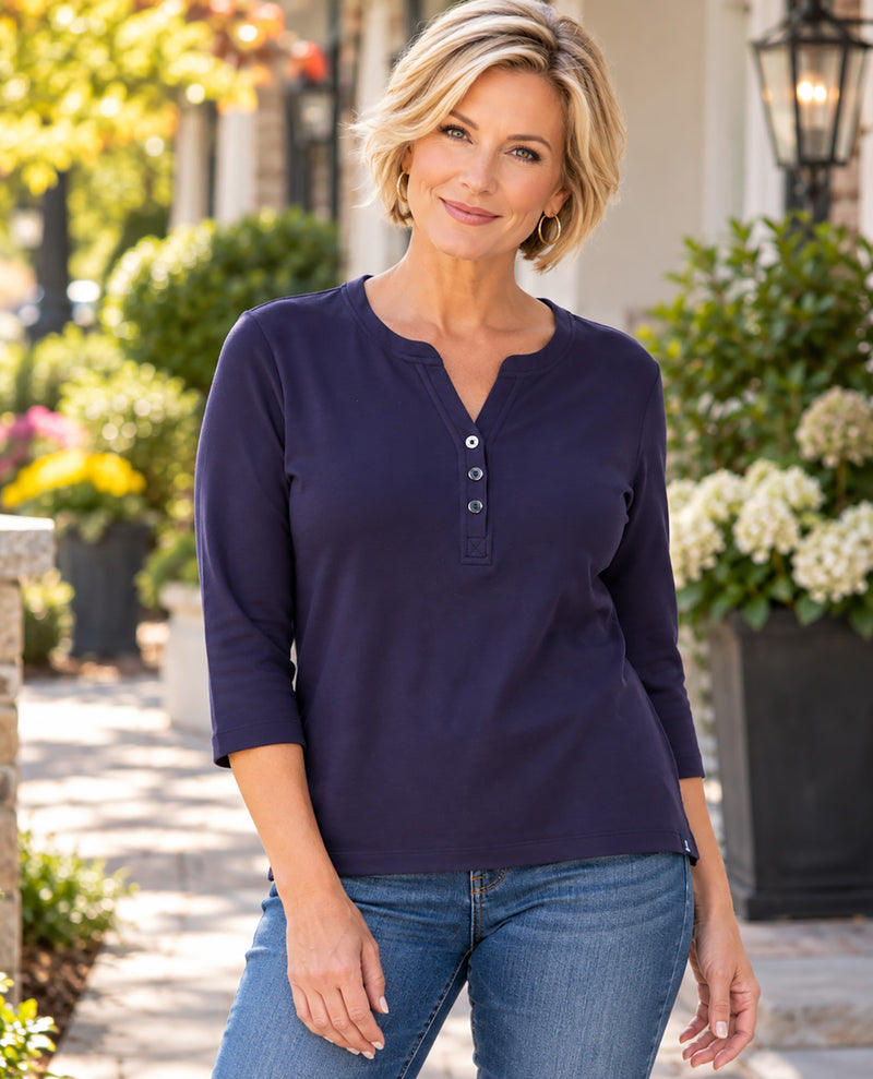 A woman wearing a navy blue henley top with three-quarter sleeves and blue jeans, standing outdoors in front of a blurred garden and house background.