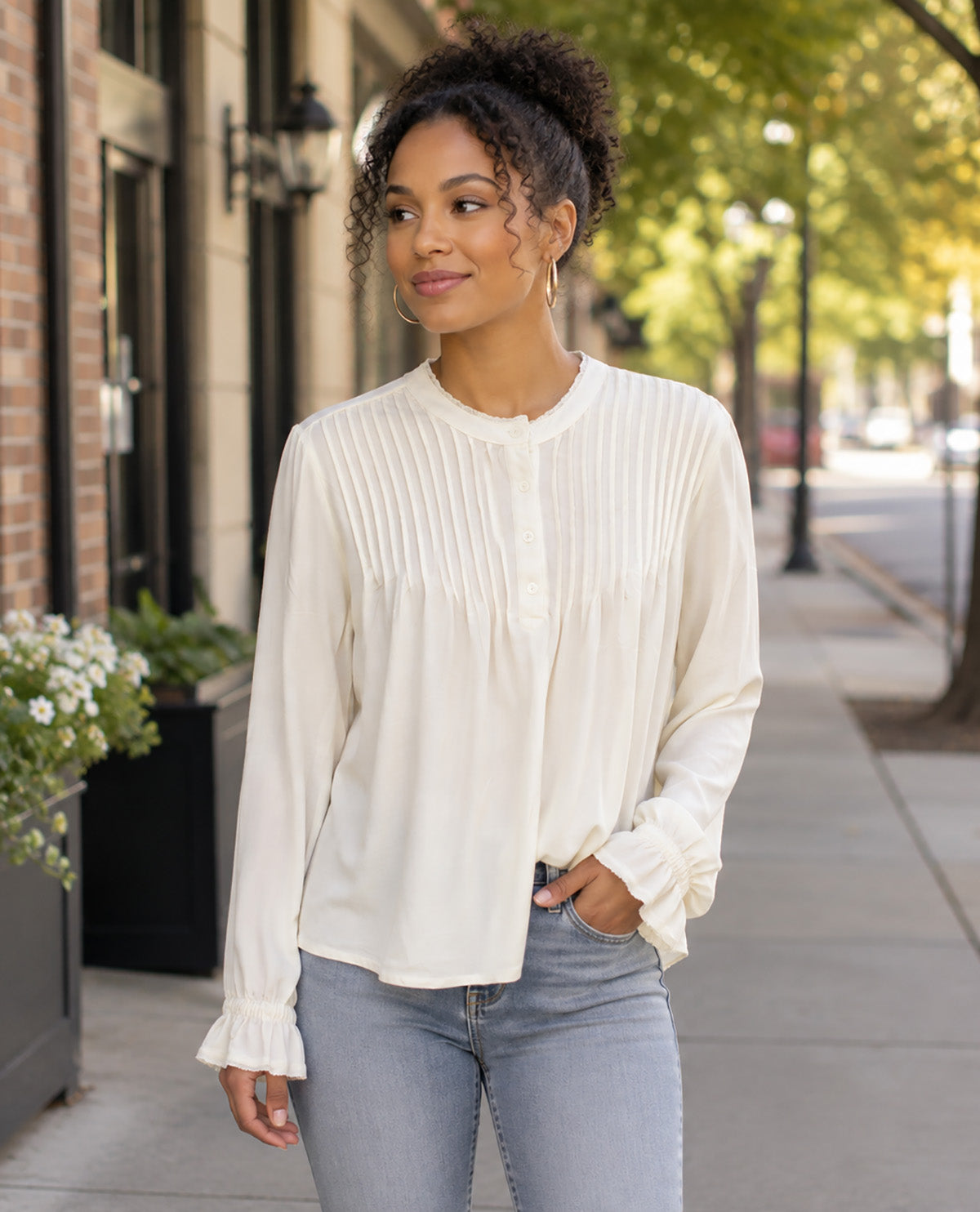 A woman wearing a cream-colored long-sleeve blouse with pleated detailing and ruffled cuffs, paired with light-wash blue jeans, standing on a city sidewalk.