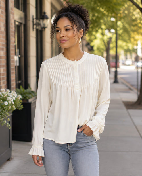 A woman wearing a cream-colored long-sleeve blouse with pleated detailing and ruffled cuffs, paired with light-wash blue jeans, standing on a city sidewalk.