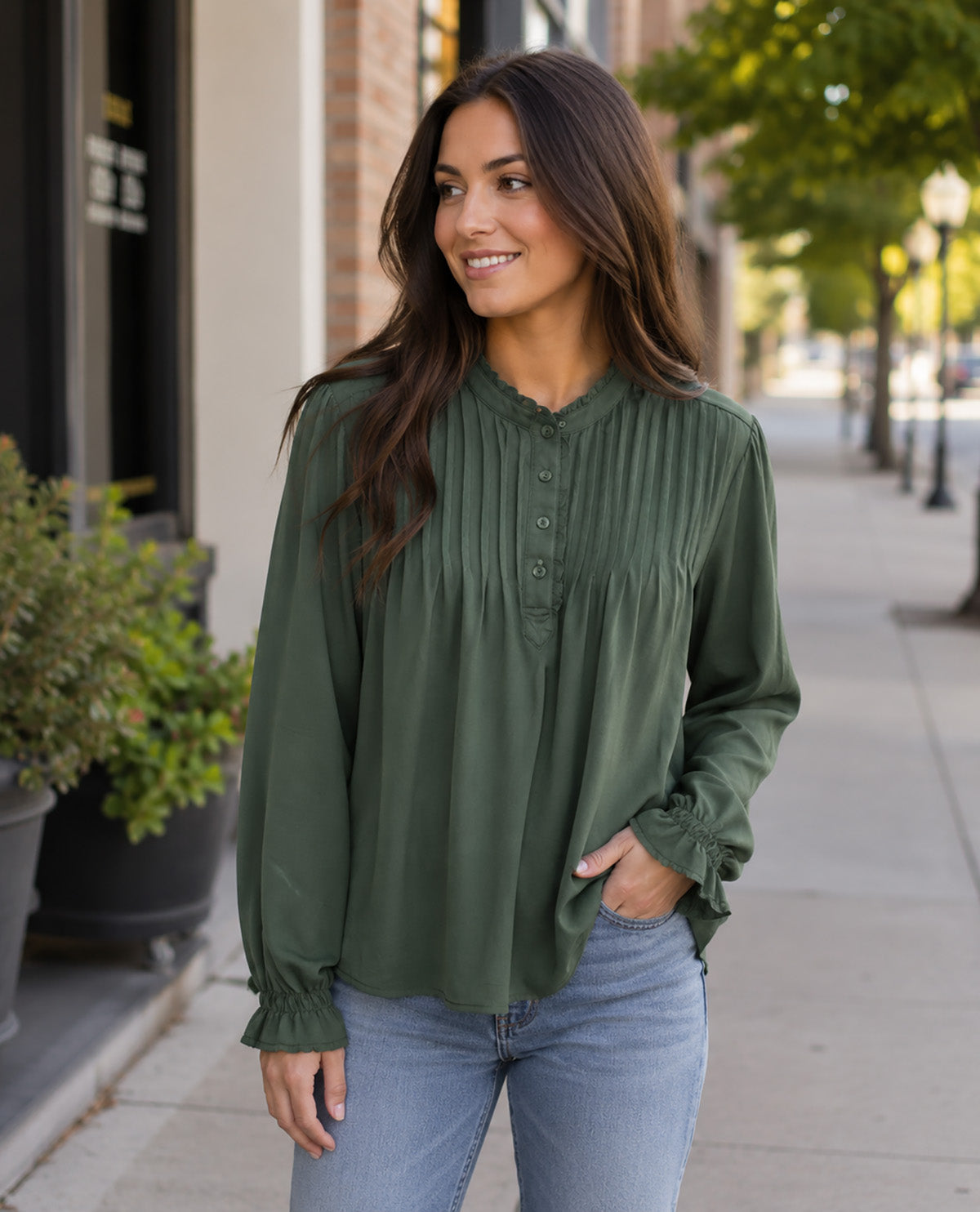 A woman wearing an olive green long-sleeve blouse with pleated detailing and ruffled cuffs, paired with light blue jeans, standing on a city sidewalk.