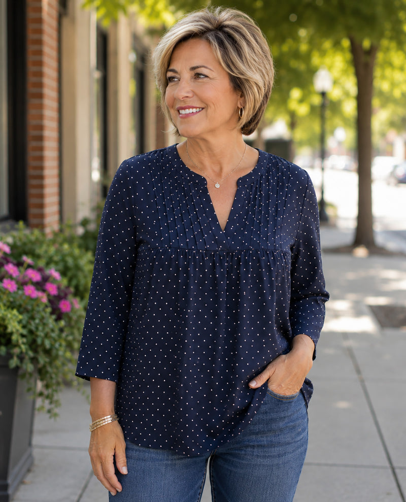 A woman with short blonde hair wearing a navy blue long-sleeve blouse with a white polka-dot pattern and blue jeans, standing on a city sidewalk.