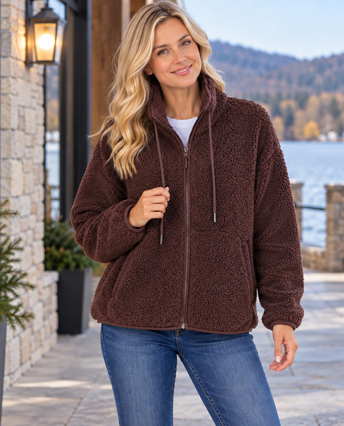 A woman wearing a brown sherpa fleece zip-up hoodie with drawstrings, styled with a white t-shirt and blue jeans, standing outdoors near a stone wall and water.