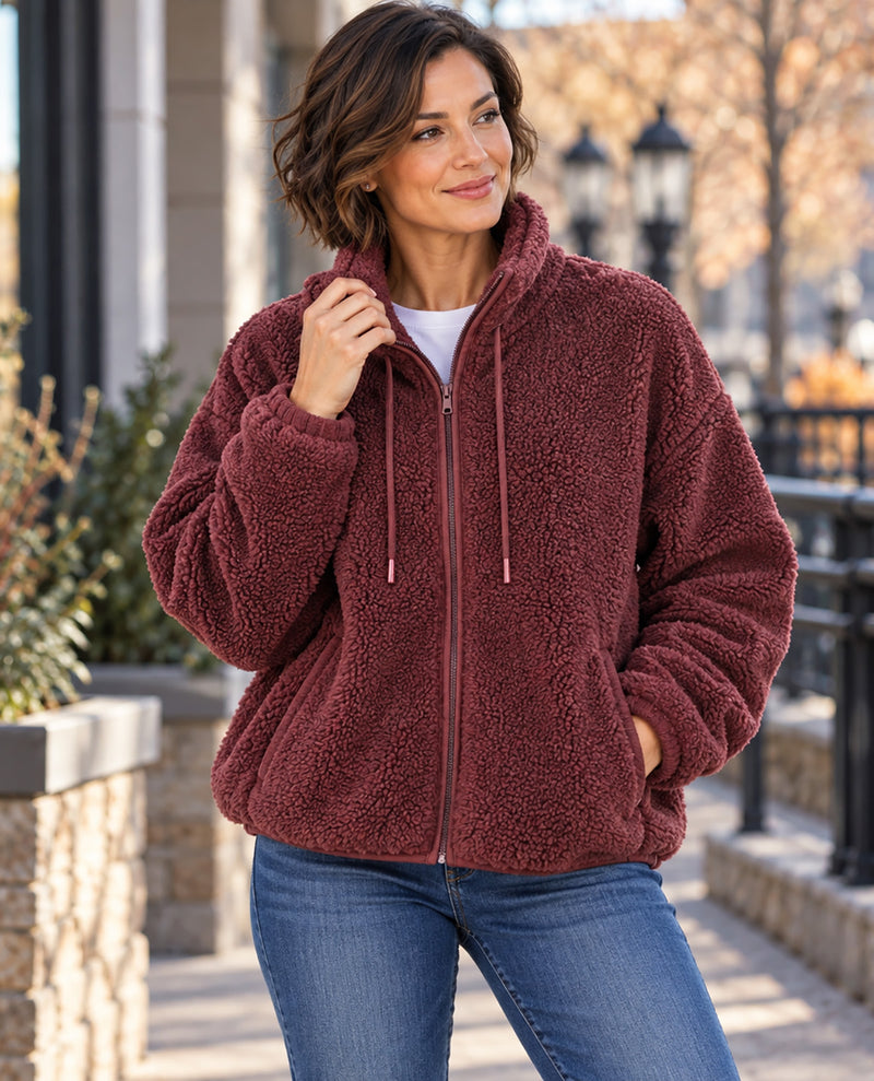 A woman wearing a plush, fleece zip-up jacket in a dusty burgundy color, paired with a white t-shirt and blue jeans, standing outdoors on a stone walkway.