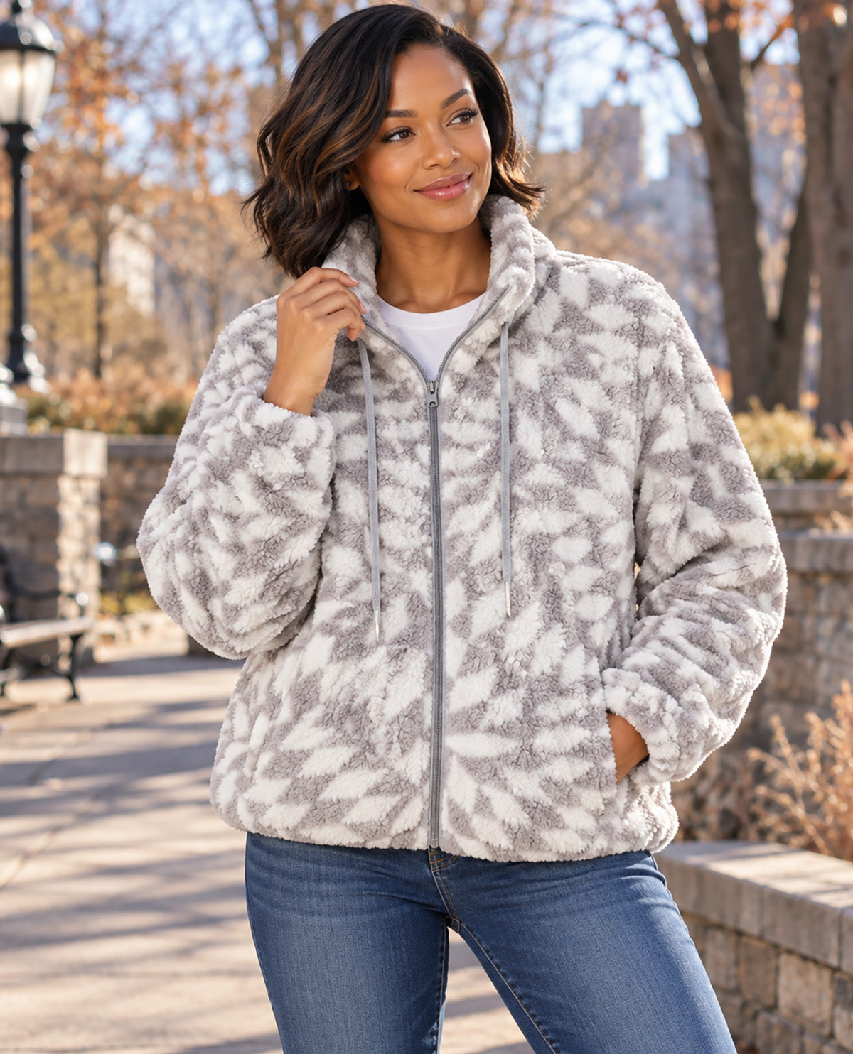 A woman wearing a plush zip-up fleece jacket with a gray and off-white abstract pattern, paired with blue jeans, standing outdoors in a park setting.