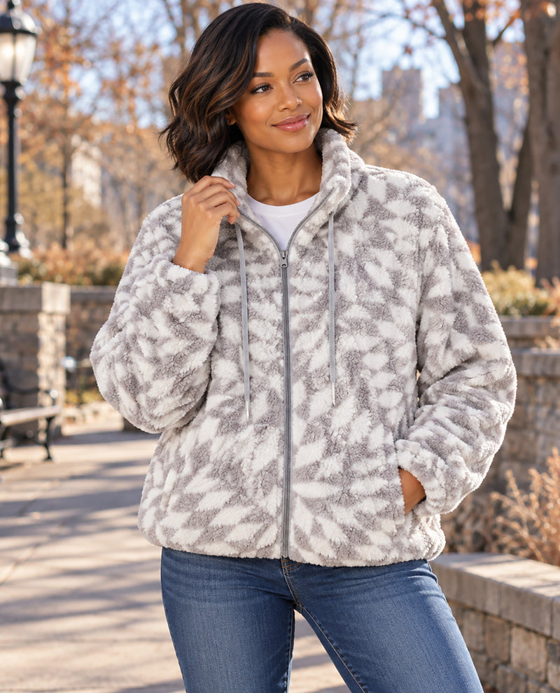 A woman wearing a plush zip-up fleece jacket with a gray and off-white abstract pattern, paired with blue jeans, standing outdoors in a park setting.