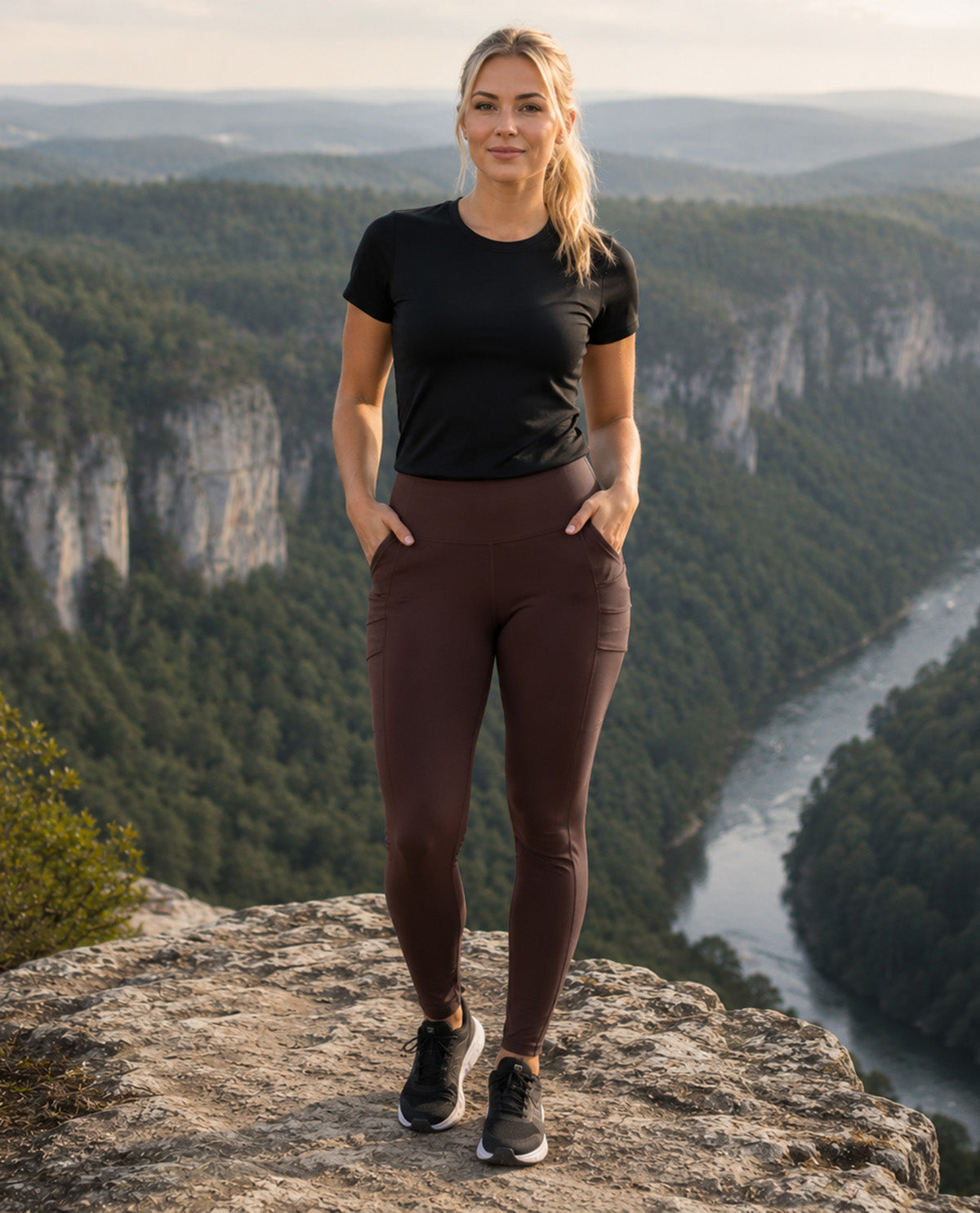 A woman stands on a rocky cliff overlooking a deep forest gorge and river, wearing a fitted black short-sleeve athletic top and dark brown high-waisted leggings with side pockets, paired with black running shoes.