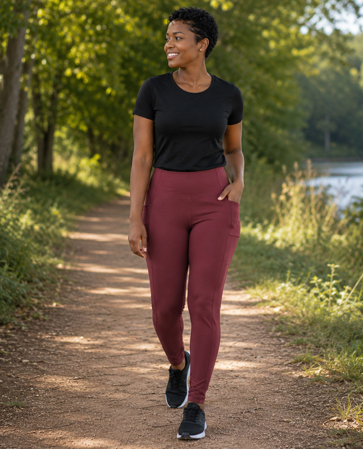 A woman walking on a sunlit dirt path outdoors, wearing a black short-sleeve athletic top and high-waisted burgundy leggings with side pockets, paired with black running shoes.