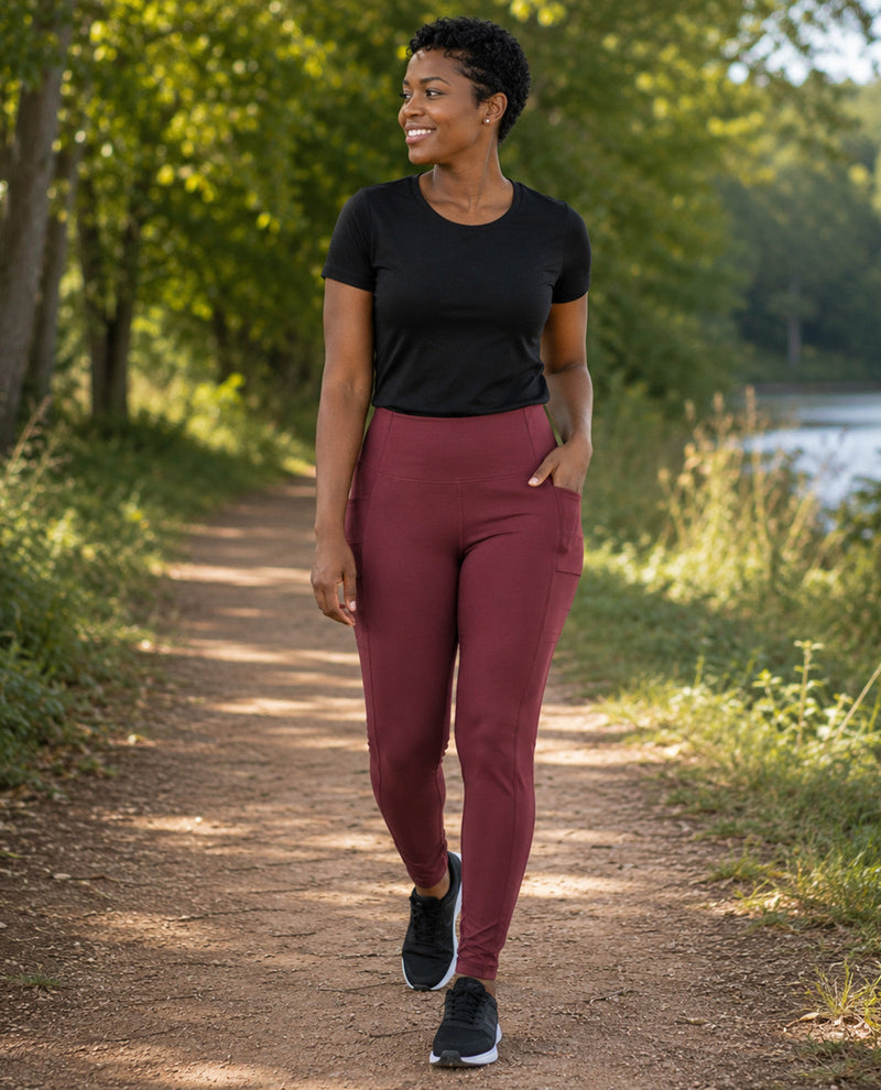 A woman walking on a sunlit dirt path outdoors, wearing a black short-sleeve athletic top and high-waisted burgundy leggings with side pockets, paired with black running shoes.