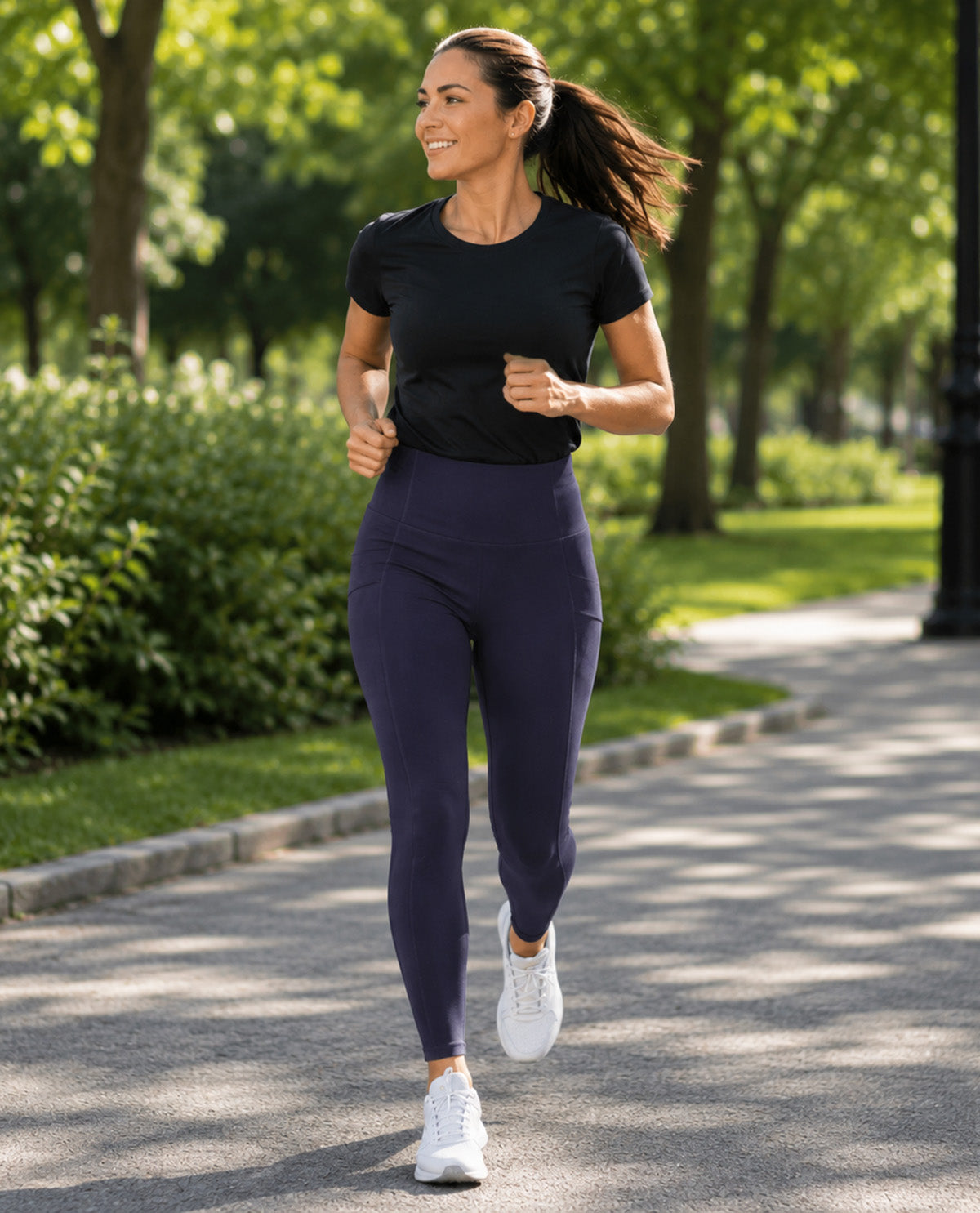 A woman jogging outdoors on a tree-lined path, wearing a fitted black short-sleeve athletic T-shirt and high-waisted navy leggings with white sneakers.