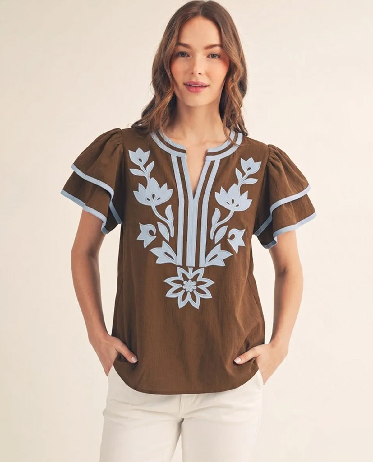 A woman modeling a brown short-sleeve blouse with light blue and white floral embroidery on the front, paired with white pants, against a plain beige background.