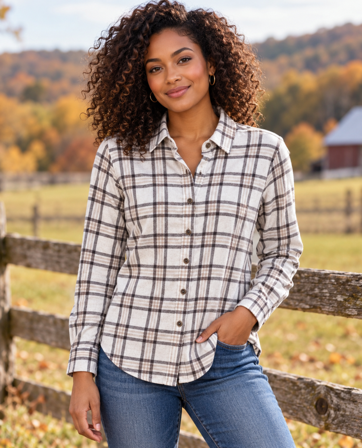 A woman with curly brown hair wearing a cream-colored long-sleeve plaid button-up shirt with brown and black lines, paired with blue jeans, standing outdoors in front of a wooden fence with an autumn landscape background.
