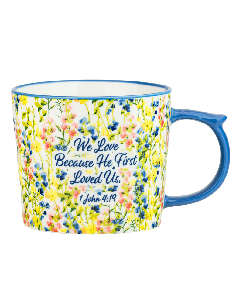 A ceramic mug with a blue handle and blue rim, decorated with a white background covered in colorful wildflowers and featuring the printed text 'We Love Because He First Loved Us. I John 4:19' in blue script.