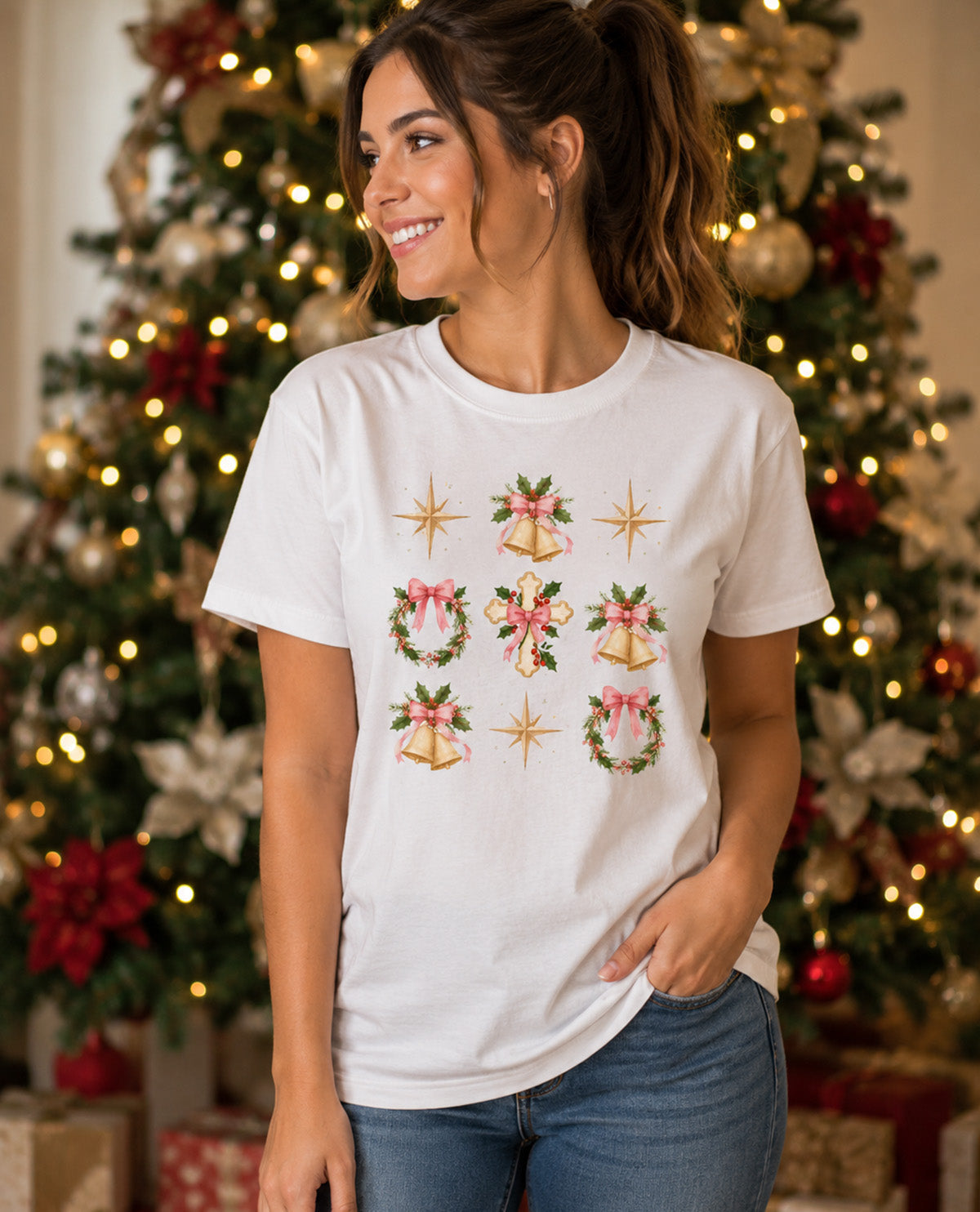 A woman wearing a white short-sleeve t-shirt with a Christmas-themed graphic print featuring bells, wreaths, and stars, posed in front of a decorated Christmas tree.