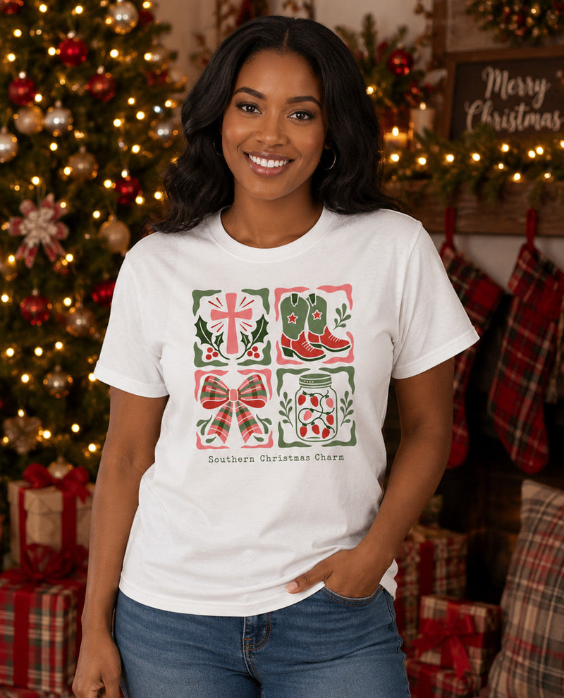 A woman wearing a white graphic t-shirt with a red and green Southern Christmas theme print, standing in front of a decorated Christmas tree and stockings.