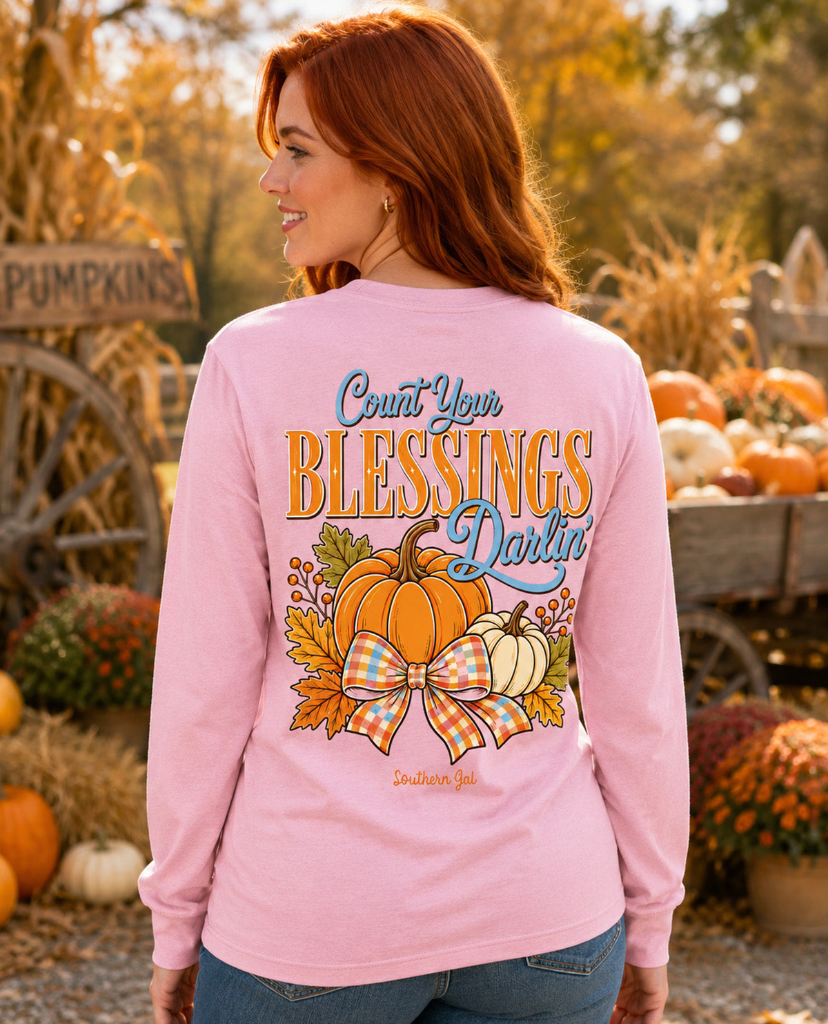 A woman with red hair is shown from the back wearing a light pink long-sleeve t-shirt with a large autumn-themed graphic on the back. The graphic features pumpkins, fall leaves, a plaid bow, and the text 'Count Your Blessings Darlin''.