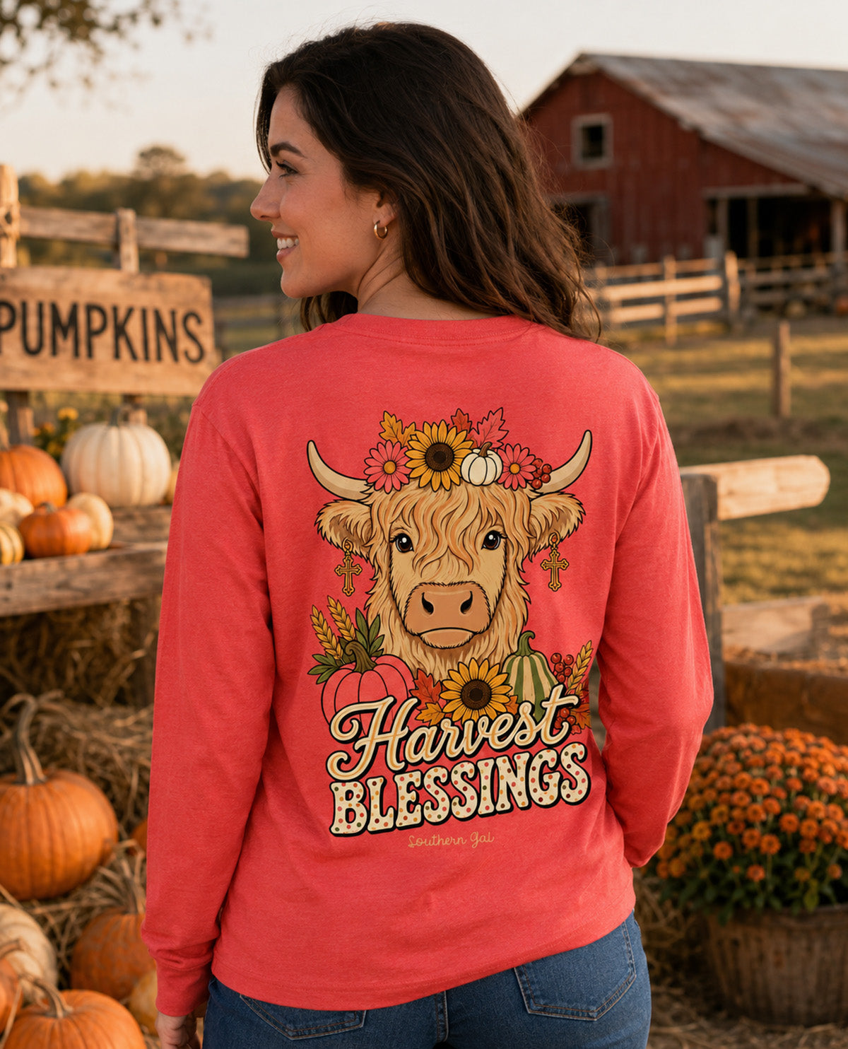 A woman wearing a coral pink long-sleeve shirt with a large autumn-themed graphic on the back, standing in a fall farm setting with pumpkins and a red barn in the background.