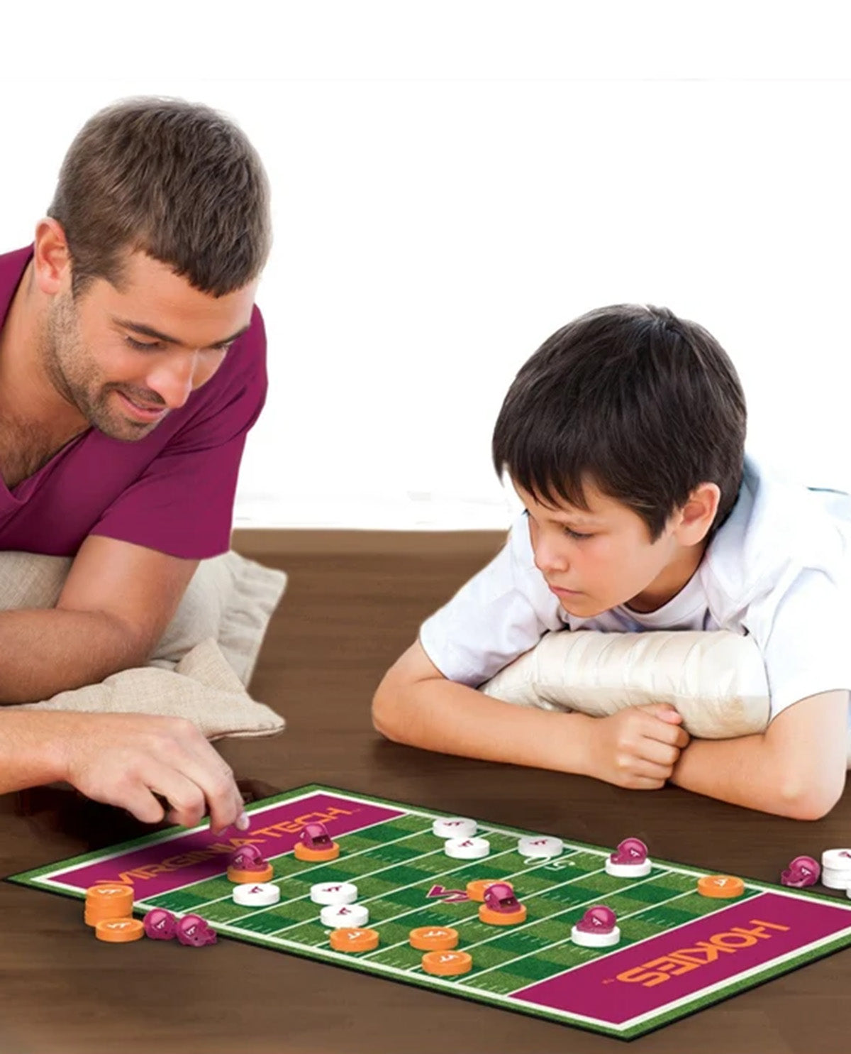 A man and a young boy are playing a football-themed board game, Hoke's, on a wooden table. The board is green with purple end zones, and the players use orange, white, and purple tokens.