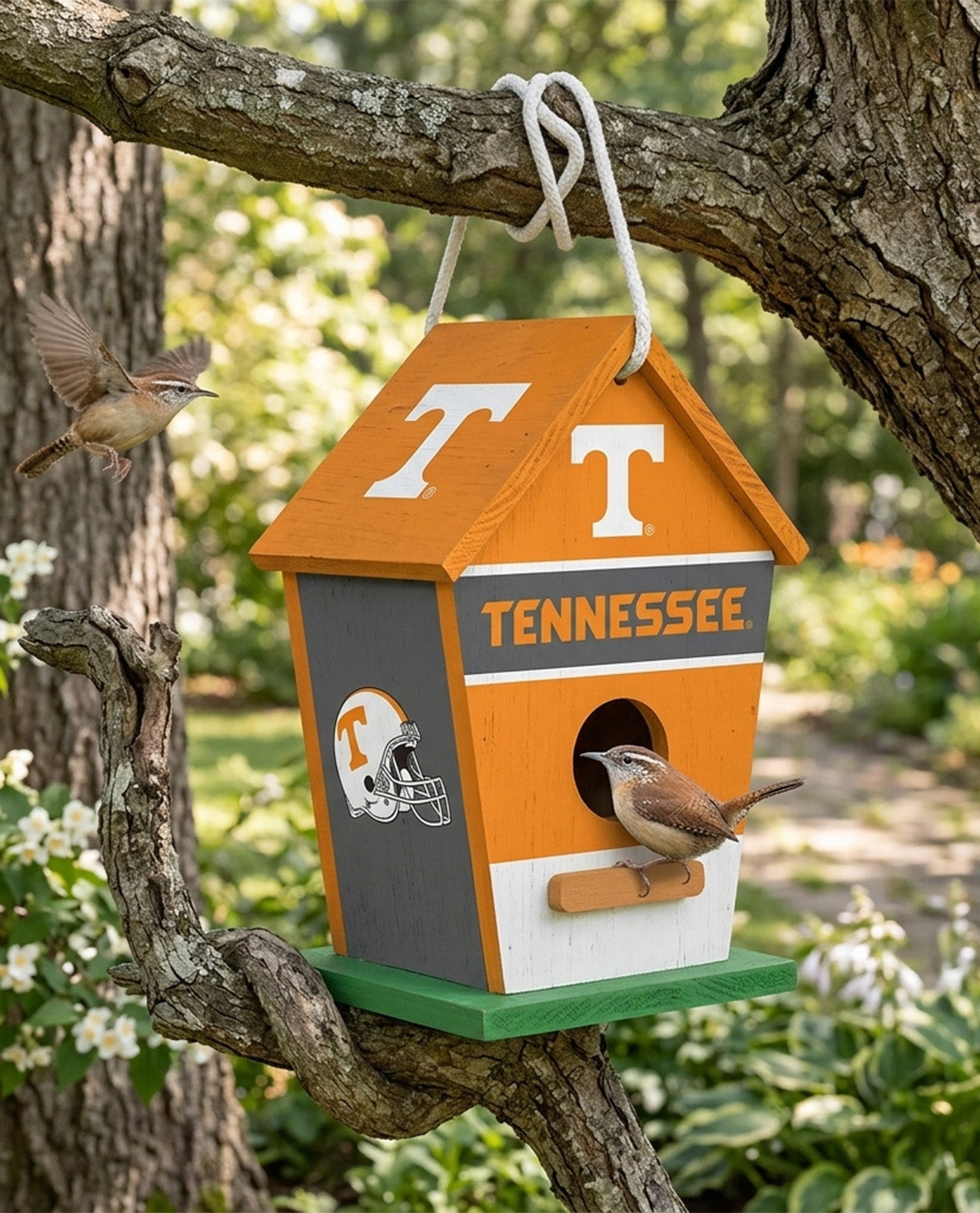 A Tennessee Volunteers themed birdhouse hanging from a tree branch outdoors. The birdhouse features an orange roof with a white 'T', a gray front panel with 'TENNESSEE' in orange, a white bottom section, a green base, and a small orange helmet graphic.