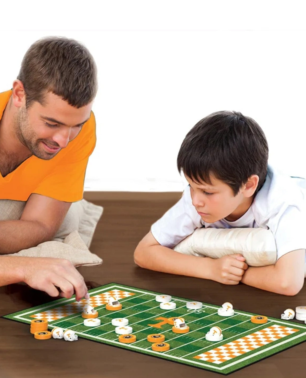 A man and a boy are playing a board game on a football-field-themed mat over a wooden table. The man wears an orange shirt and is moving a piece, while the boy, with a cast on his arm, watches closely.