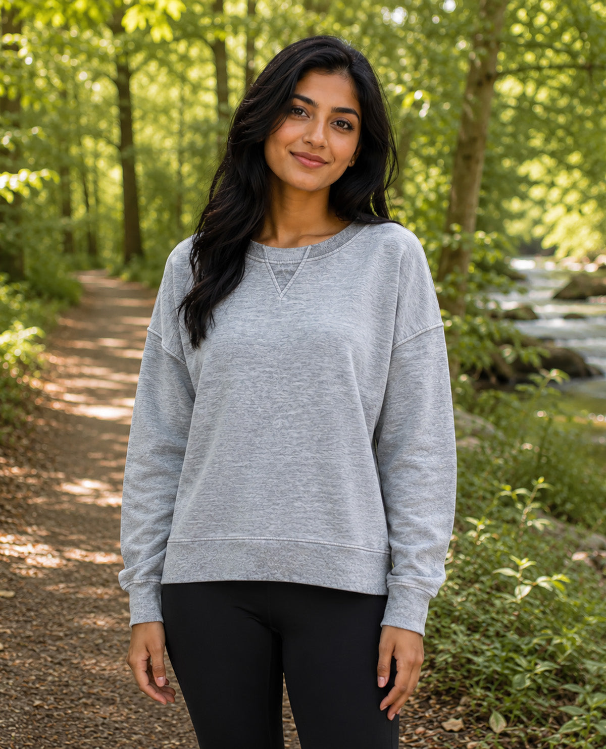 A woman wearing a light gray heather crewneck sweatshirt and black leggings standing on a forest path with trees and a stream in the background.