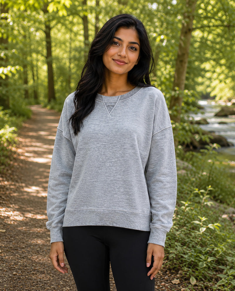 A woman wearing a light gray heather crewneck sweatshirt and black leggings standing on a forest path with trees and a stream in the background.