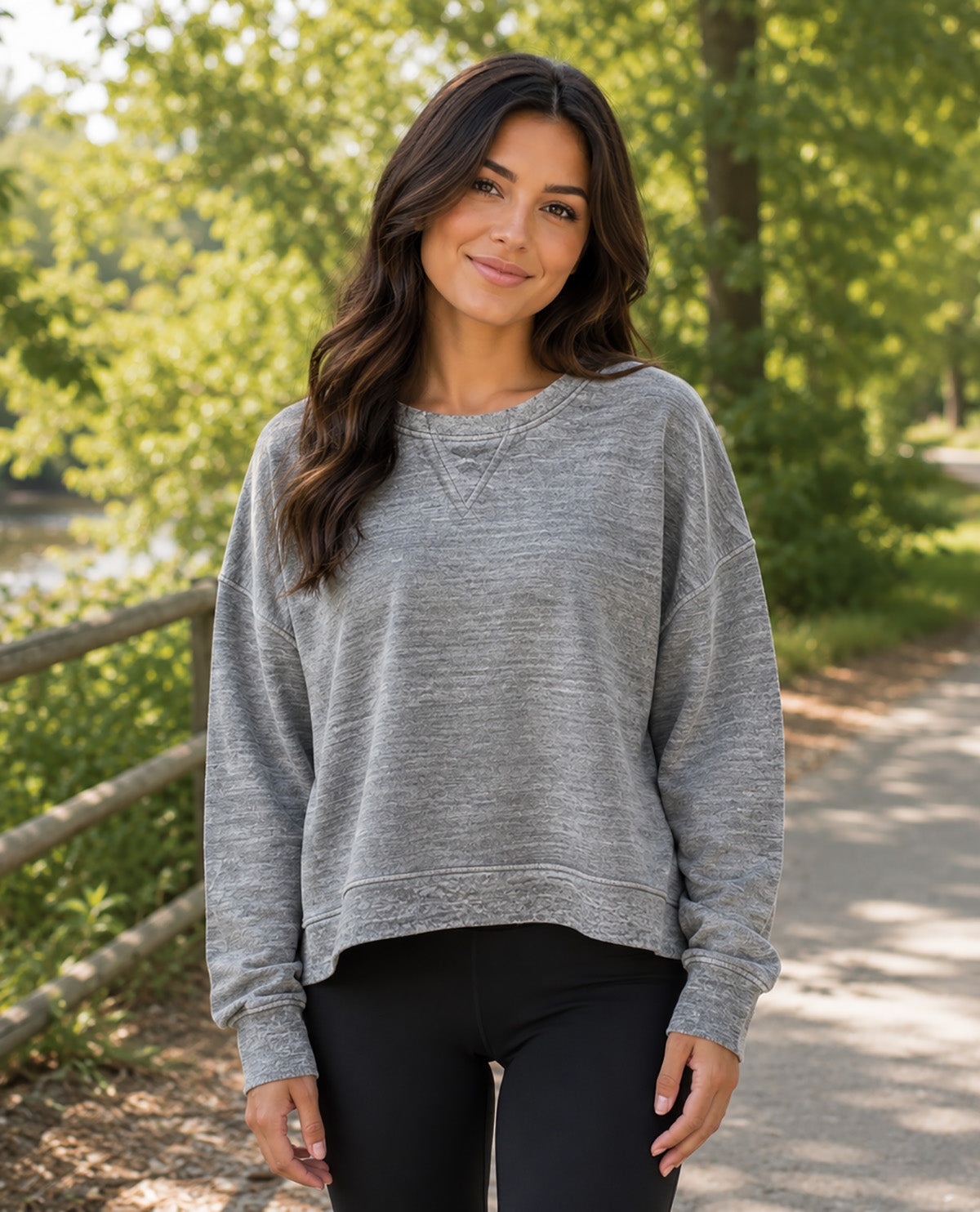A woman wearing a heather gray long-sleeve crewneck sweatshirt and black pants, standing outdoors on a sunlit path with trees and a fence in the background.
