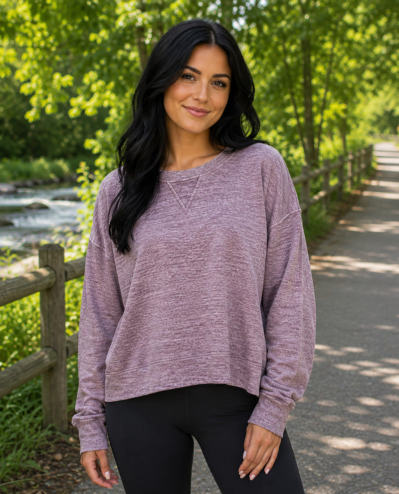 A woman with long black hair stands on an outdoor paved path wearing a heathered dusty mauve long-sleeve crewneck sweatshirt and black leggings, with a wooden fence and green trees in the background.