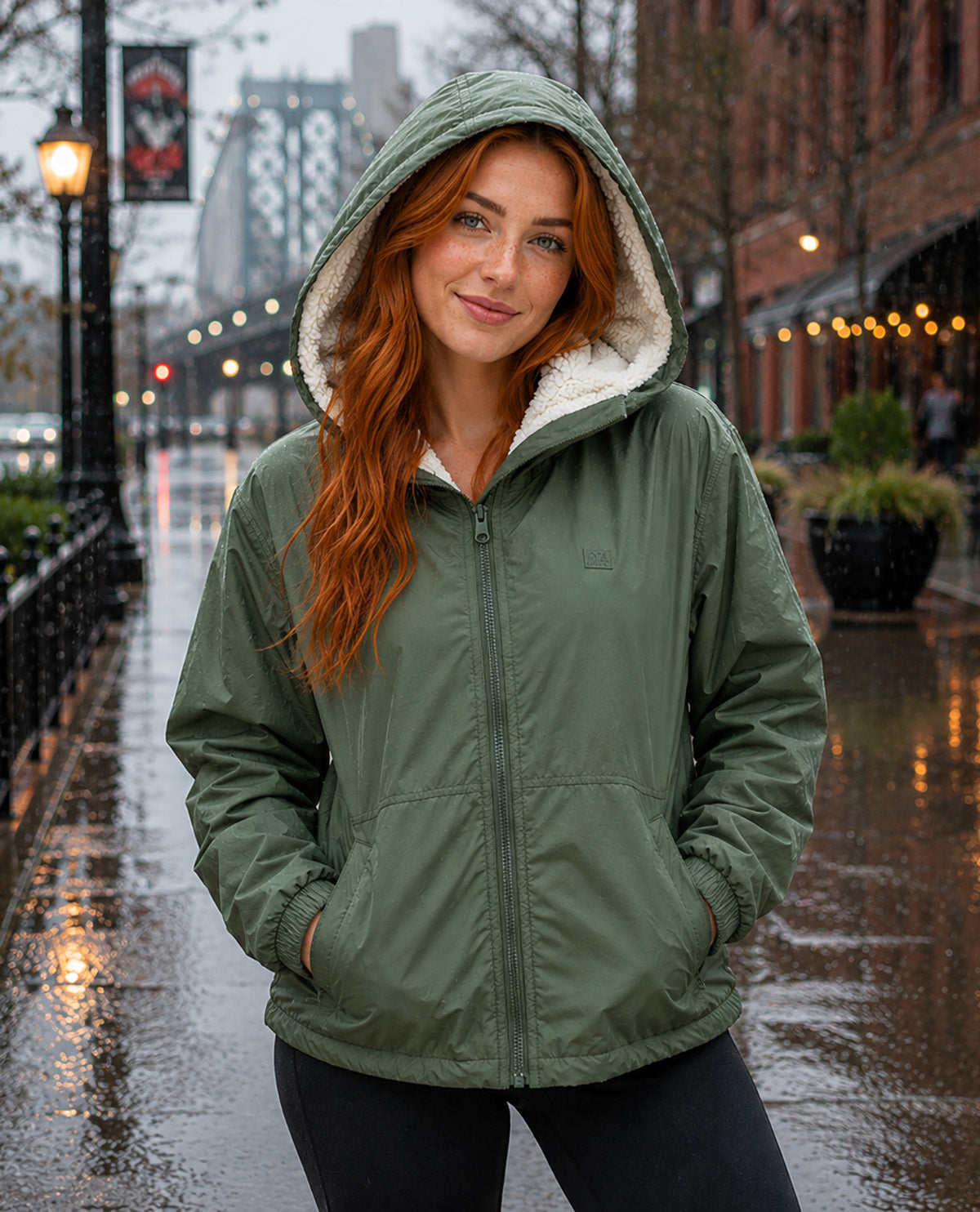 A woman with long red hair wearing an olive green hooded jacket with a white sherpa lining, standing on a wet city street.