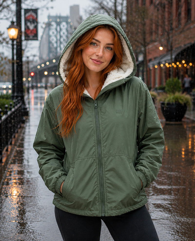 A woman with long red hair wearing an olive green hooded jacket with a white sherpa lining, standing on a wet city street.