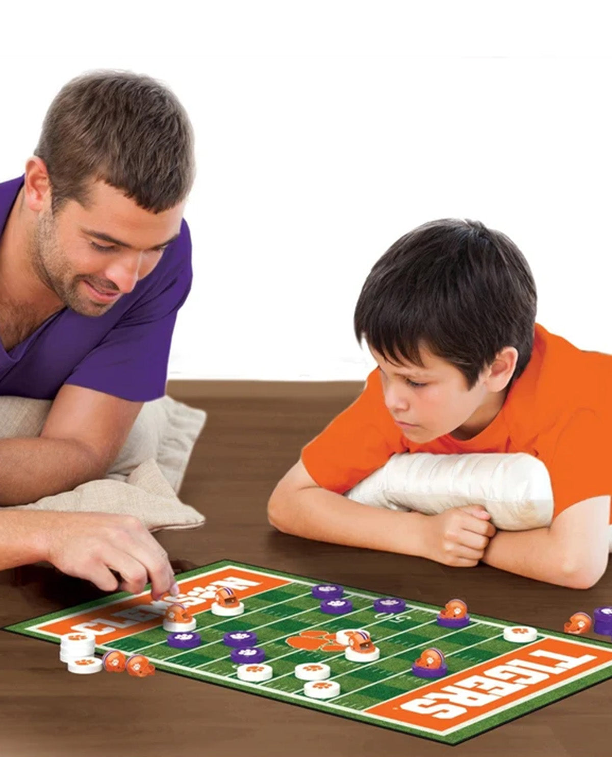 A man and a young boy are playing a football-themed board game on a wooden table. The board is green with orange and white sections, and the boy wears an orange shirt with a white cast on his arm.