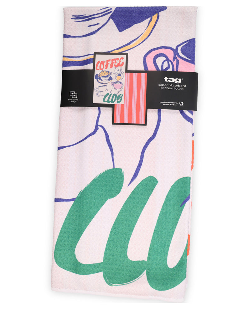 A light pink waffle-weave kitchen towel with a colorful abstract graphic print, featuring a black paper band with the brand 'tag' and a 'COFFEE CLUB' graphic.