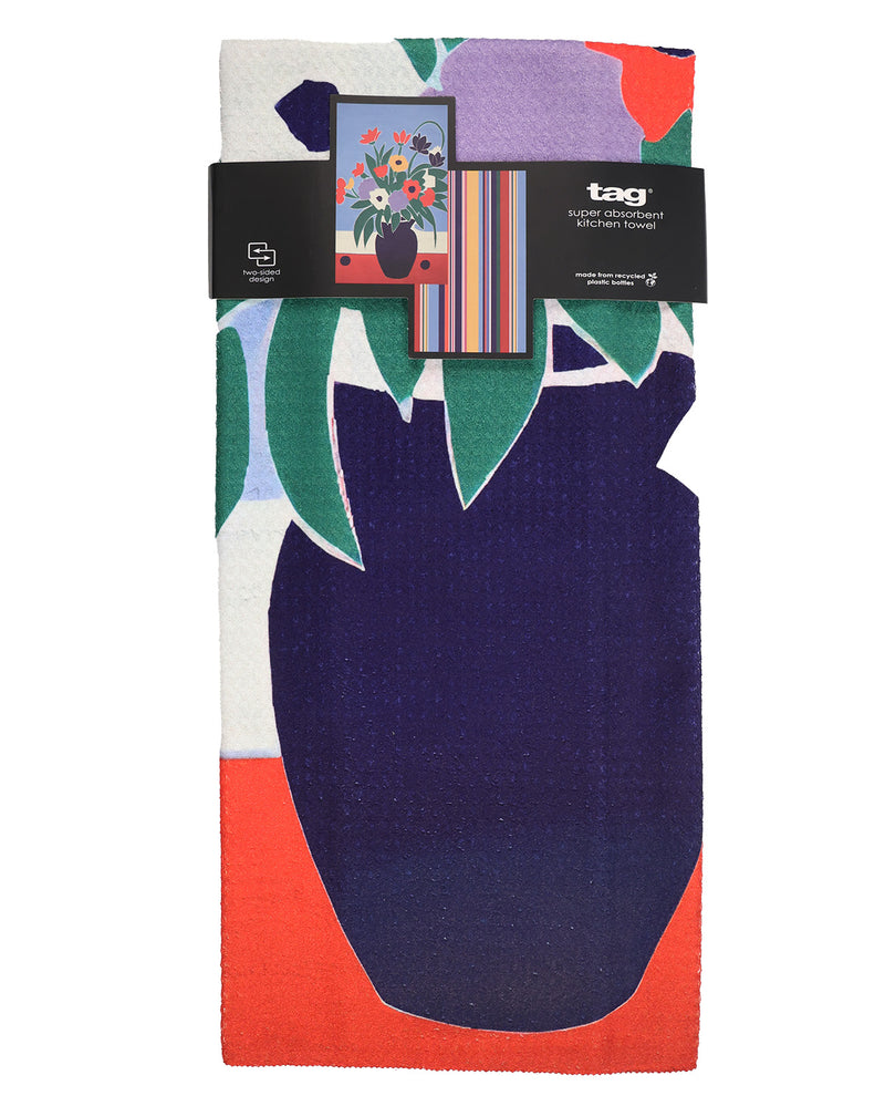 A folded cotton towel by tag featuring a colorful abstract floral and leaf pattern with a large dark navy shape, green leaves, purple and orange sections, and a black paper band label wrapped around the top.