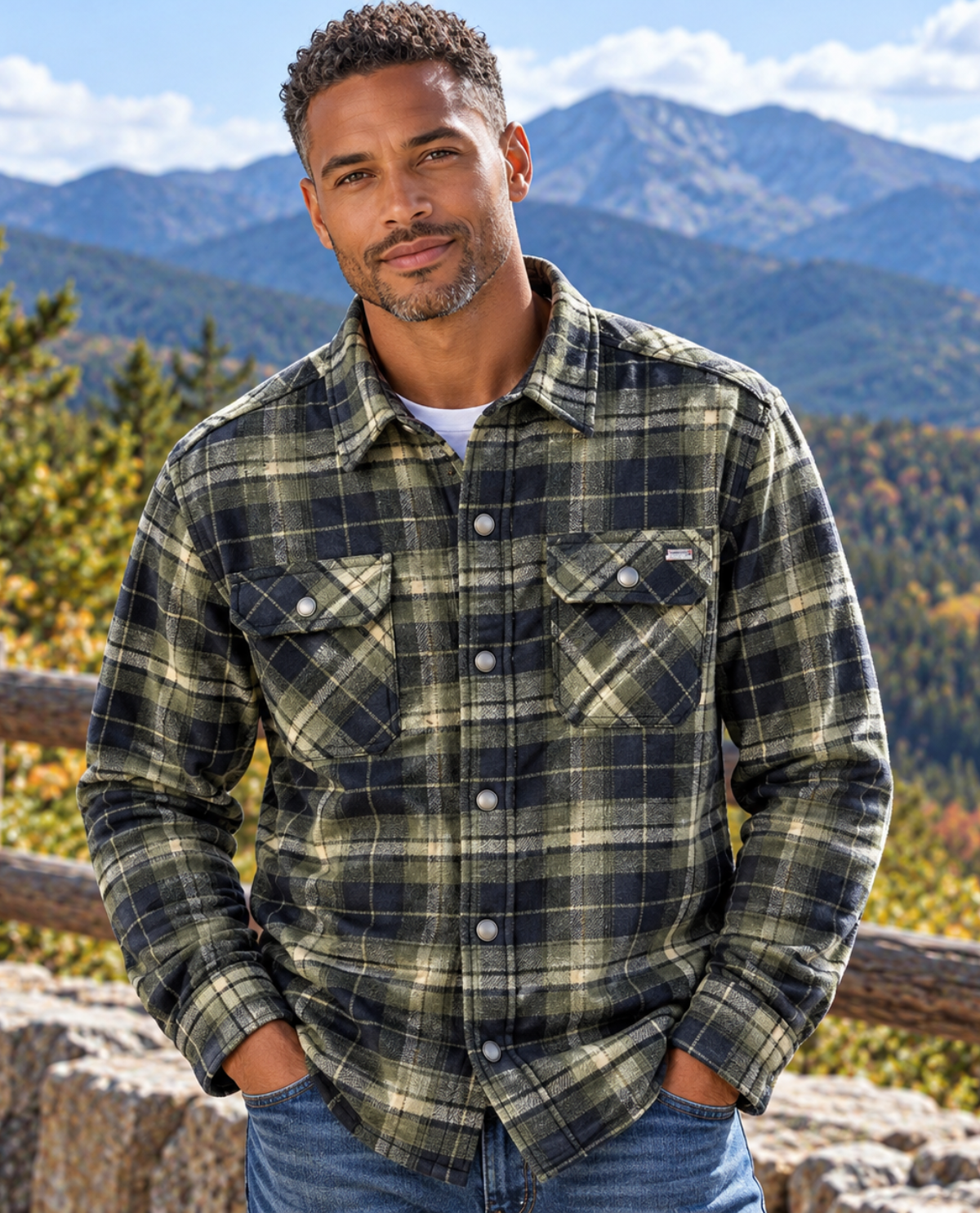 A man wearing a green and black plaid flannel shirt jacket with blue jeans, standing outdoors in front of a mountain landscape.