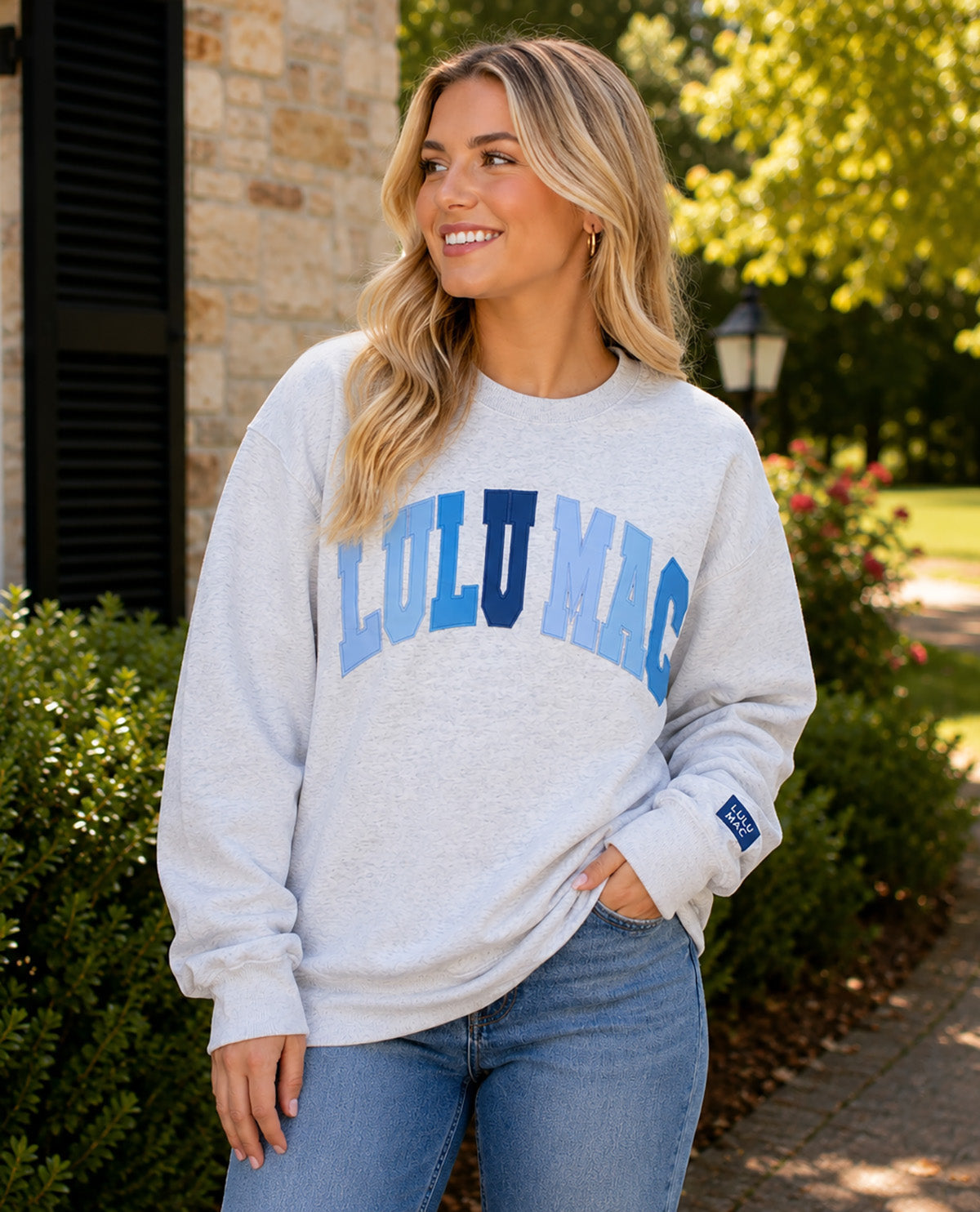 A woman wearing a light heather gray sweatshirt with 'LULU MAC' printed across the chest in blue and navy, paired with blue jeans, standing outdoors in front of a stone building and greenery.